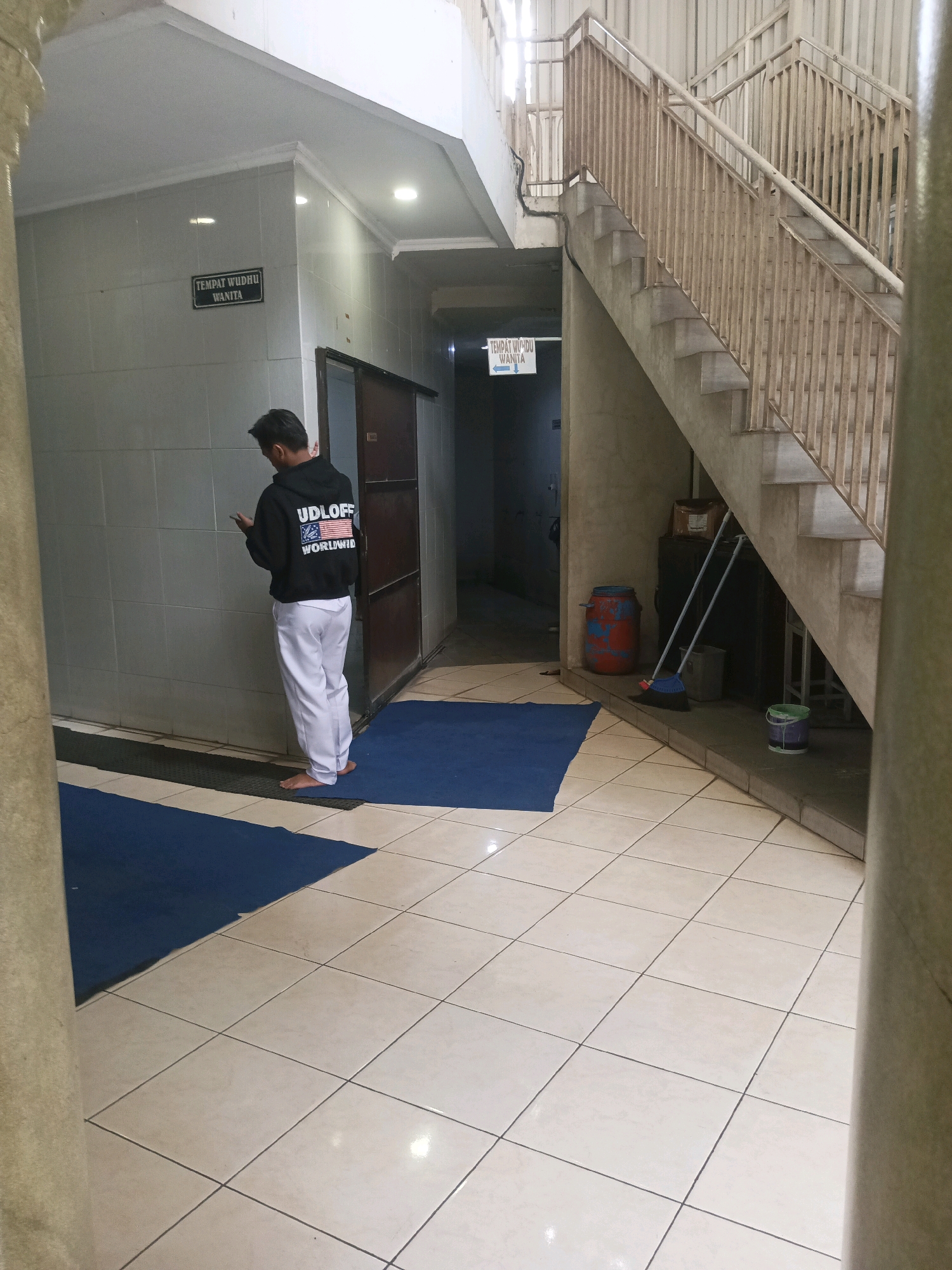 The image depicts a scene at a public facility, likely a mosque or a prayer hall, in South Tangerang, Indonesia. In the foreground, a young man wearing a black hoodie with "UDLOFF WORLDWIDE" and an American flag design on the back, and white trousers, stands barefoot on a blue mat, looking at his phone. He is positioned in front of a tiled wall. To his left, a sign on the tiled wall reads "TEMPAT WUDHU WANITA," indicating a women's ablution area. A dark brown door is visible to his right.
The environment features beige tiled flooring throughout. To the right of the man, a staircase with a light-colored metal railing ascends. At the base of the stairs, a red and blue barrel, a broom with a long handle, and a green bucket are visible. The overall setting appears to be an indoor, covered walkway or corridor leading to facilities.