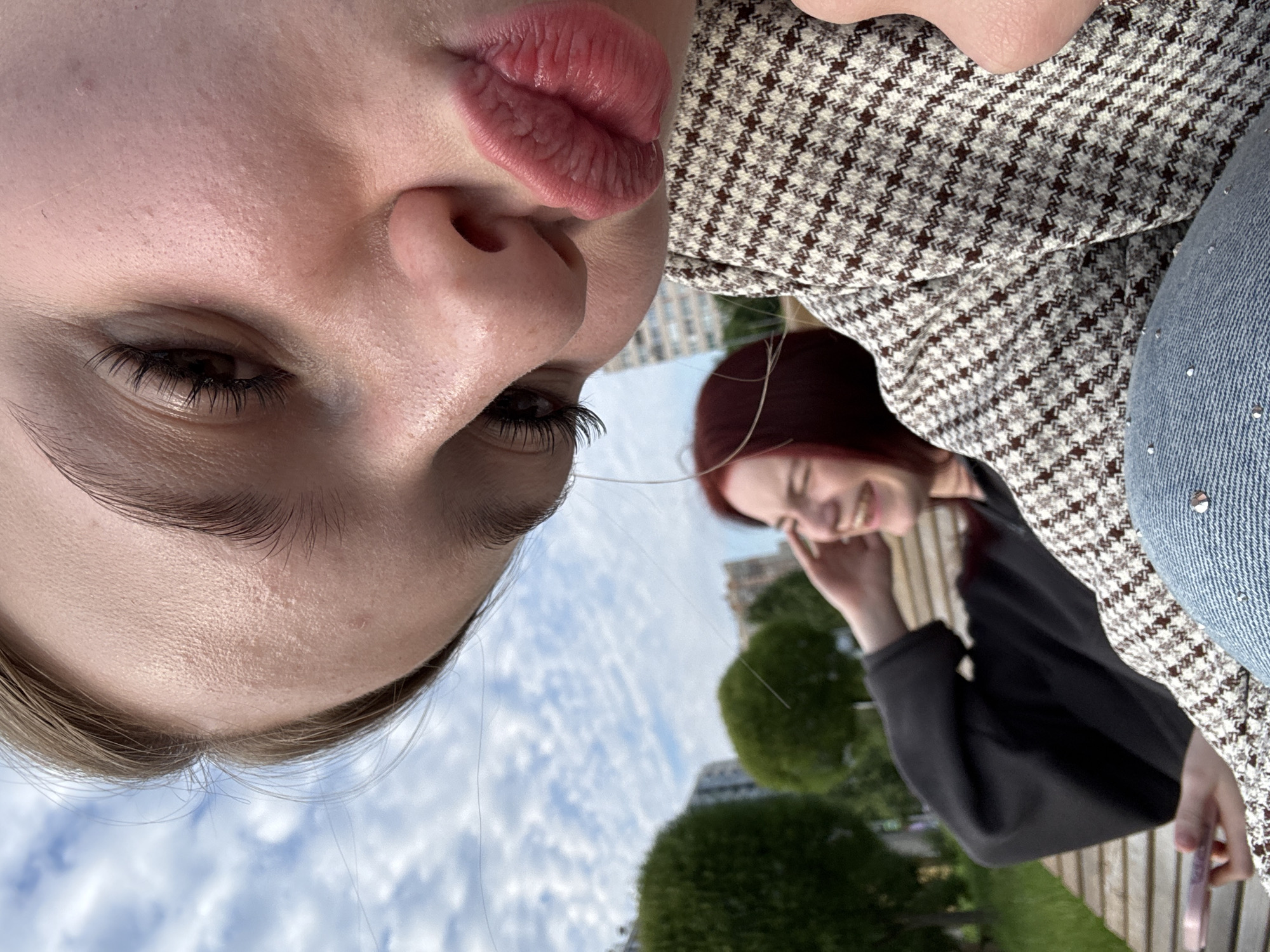 Two young women are posing for a selfie in an outdoor urban park area. The woman in the foreground is close to the camera, making a kissing face with her lips pursed. Her face is partially obscured by the camera angle. She is wearing a patterned jacket and blue jeans. Behind her, slightly out of focus, the second woman is visible sitting on a wooden bench, laughing with her hand near her face. She is wearing a black sweatshirt. The background shows green trees, buildings, and a cloudy sky. The scene is consistent with a park in Sinyavino, Russia, given the architectural style of the buildings and the overall urban park setting.
