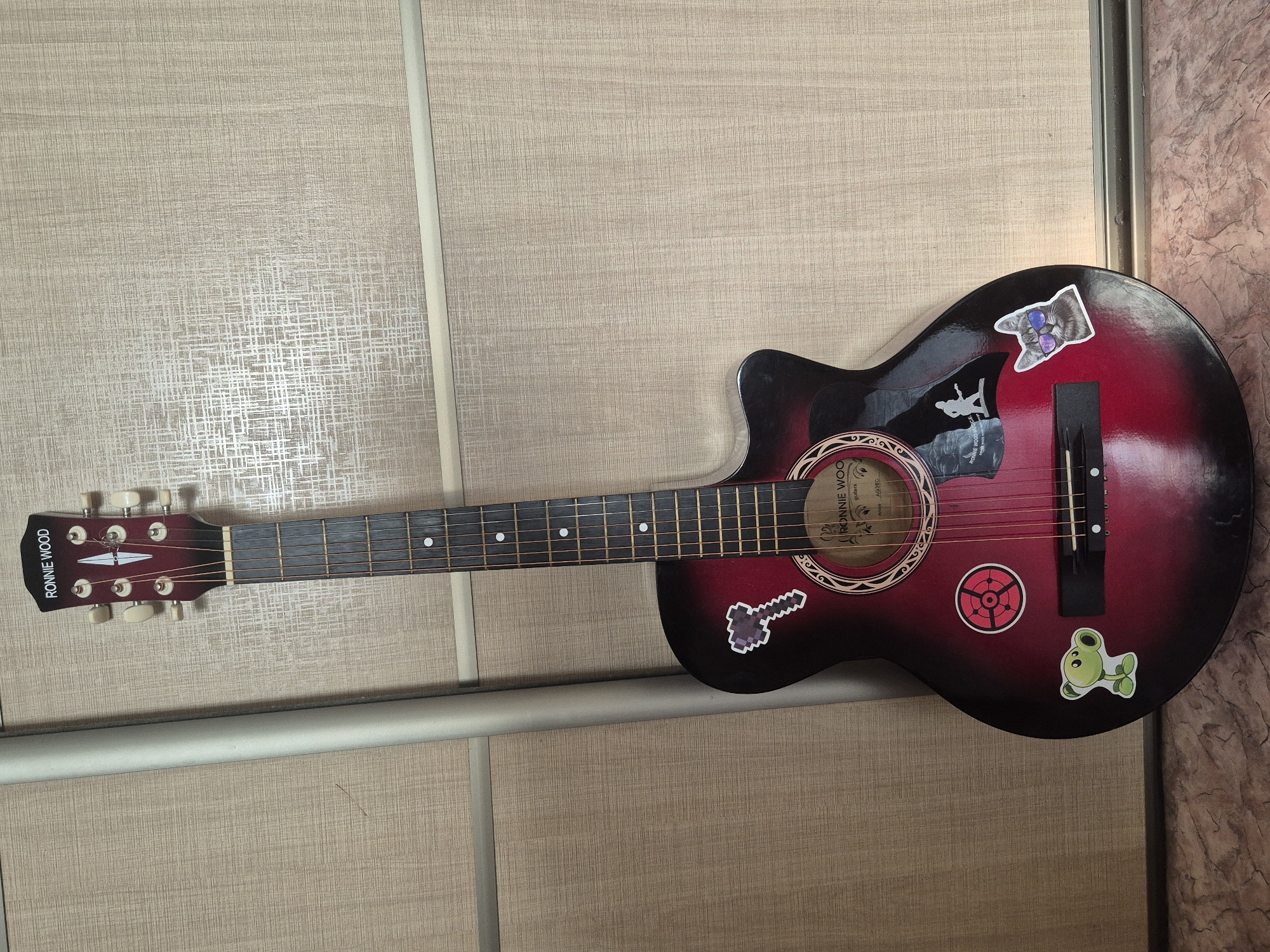 An acoustic guitar, branded "RONNIE WOOD," stands leaning against a textured, light-colored wall, likely a closet door. The guitar has a gradient finish, transitioning from black at the top to a deep red at the body. It is adorned with several stickers: a pixelated hammer on the upper left bout, a stylized target with a red circle and crosshairs on the lower left bout, a cartoon pea-shooter plant from "Plants vs. Zombies" on the lower left, a silhouette of a guitarist on the upper right bout, and a cat wearing sunglasses on the lower right bout. The guitar's soundhole has a circular decal with branding. The floor beneath the guitar is a patterned surface with varying shades of brown and pink. The image was taken indoors, possibly in a residential setting in Kokshetau, Kazakhstan.
