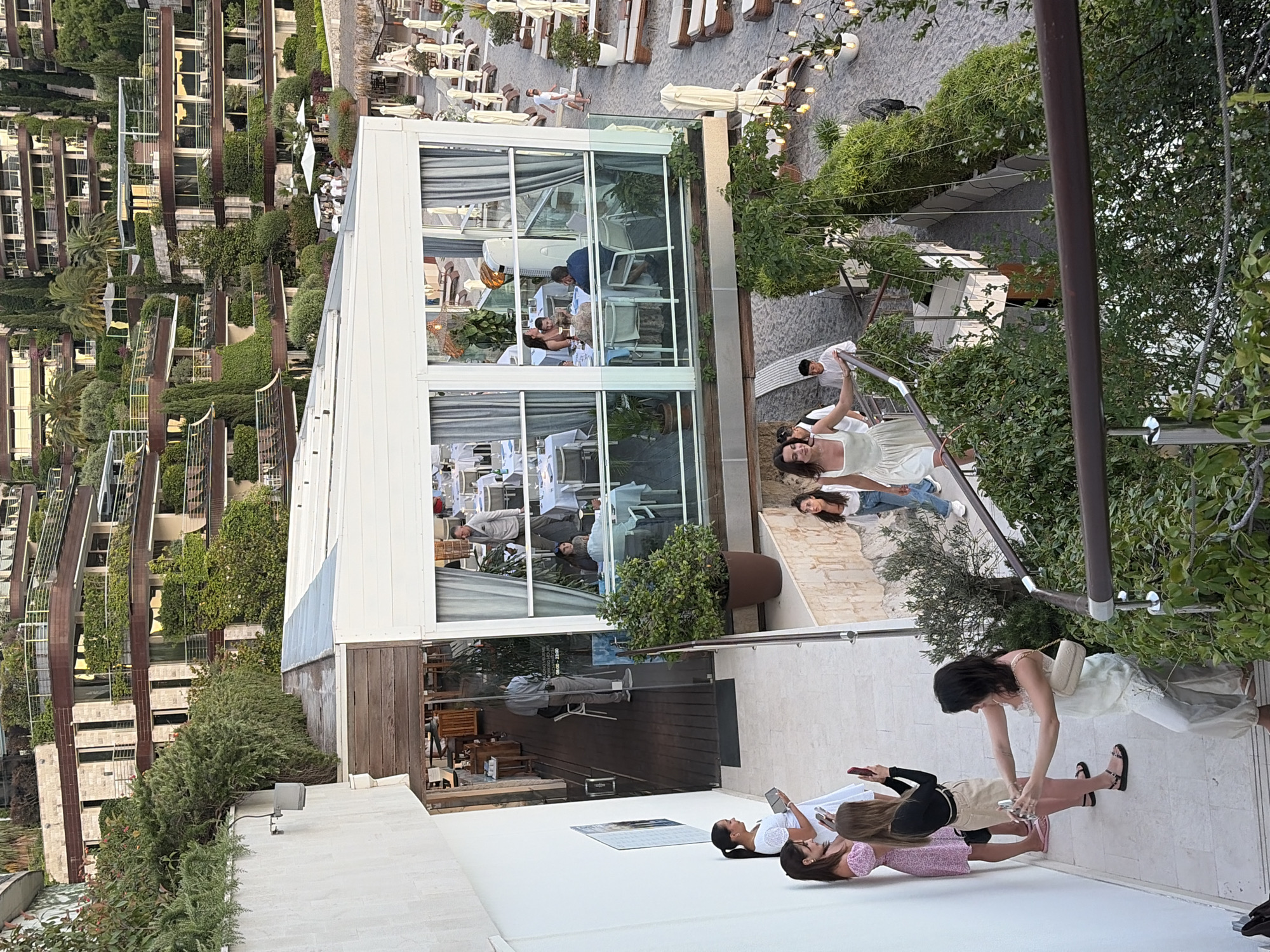 The image captures a scene at a resort or hotel complex in Budva, Montenegro, featuring a tiered, multi-story building with extensive greenery cascading down its facade. In the foreground, several women are descending a staircase. One woman in a white dress is taking a selfie with a camera, while others are either looking at their phones or posing. To the left, a white wall has a framed poster. Above the staircase, a modern, glass-enclosed dining area with tables set for meals is visible, with diners seated inside and staff moving around. Beyond the dining area, a beach with lounge chairs and umbrellas is apparent. The architecture suggests a contemporary resort design, with balconies and terraces integrated into the hillside. The overall ambiance is that of a lively vacation spot.