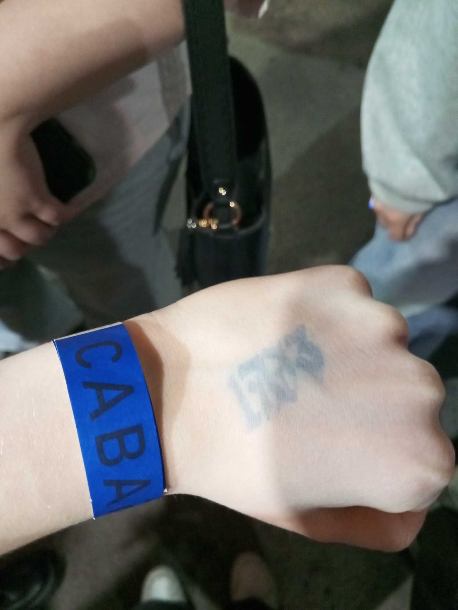 The scene depicts a close-up of a person's left forearm and hand, illuminated by dim lighting, set within the city of Saint Petersburg, Russia. A blue wristband, approximately one inch wide, encircles the wrist, displaying the black capital letters "CABA". The dorsal surface of the hand features a faded, dark grey or black tattoo, the specific design of which is indistinct due to blur and fading. In the background, other individuals are partially visible. To the left, a person's upper arm, wearing a light-colored short sleeve, holds a dark-colored smartphone. To the right, another individual's torso and right arm are visible, clad in a light grey long-sleeved top. A black shoulder bag with gold-colored metallic rings is positioned behind the primary subject's arm. The floor or ground surface beneath the subjects is dark, with the toe of a white shoe visible in the lower right corner. The presence of the "CABA" wristband indicates attendance at an event or establishment in Saint Petersburg.
