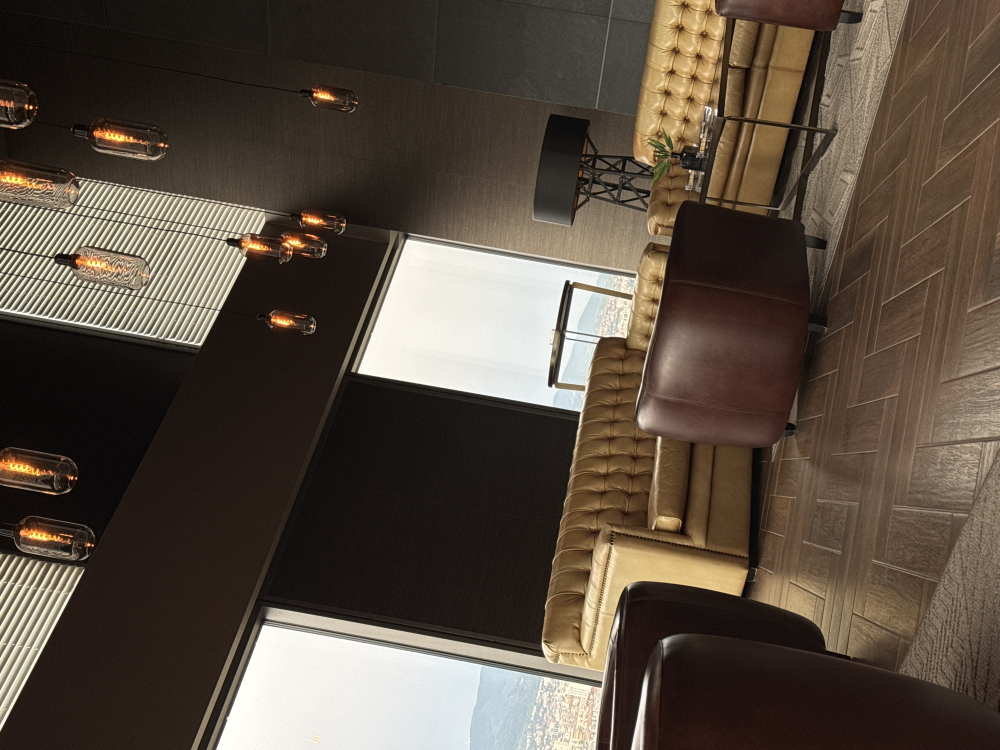 This image depicts a modern, luxurious interior space, likely a hotel lounge or reception area, situated high up in a building in Sofia, Bulgaria. Large windows offer a panoramic view of the city and distant mountains, suggesting an elevated position.
The foreground is dominated by plush, tufted seating arrangements. Two large, dark brown leather armchairs are prominently featured on the left and right. Between them, a tan, tufted sofa with a classic Chesterfield design is visible. A smaller, round, dark brown ottoman or accent chair is positioned in front of the sofa. A low, modern coffee table with a glass top and dark metal legs sits in front of the sofa, holding a small potted plant and what appear to be coasters.
The flooring is a dark wood with a herringbone pattern, and a neutral-toned rug is partially visible beneath the seating area.
The lighting is a key feature of the space, with multiple Edison-style pendant lights hanging from the ceiling. These lights, with visible filament bulbs, cast a warm, amber glow. The ceiling itself is a mix of dark panels and a large area with white blinds, through which sunlight or ambient light filters.
In the background, a tall, industrial-style floor lamp with a dark rectangular shade stands near the sofa on the right. The wall behind the lamp has a textured, dark finish. A thin, tall, dark framed object, possibly a cabinet or part of the building's structure, is also visible.
The overall impression is one of sophisticated comfort and contemporary design, with a strong emphasis on ambient lighting and luxurious materials. The view of Sofia from this vantage point is a significant element, implying the location is a premium establishment.