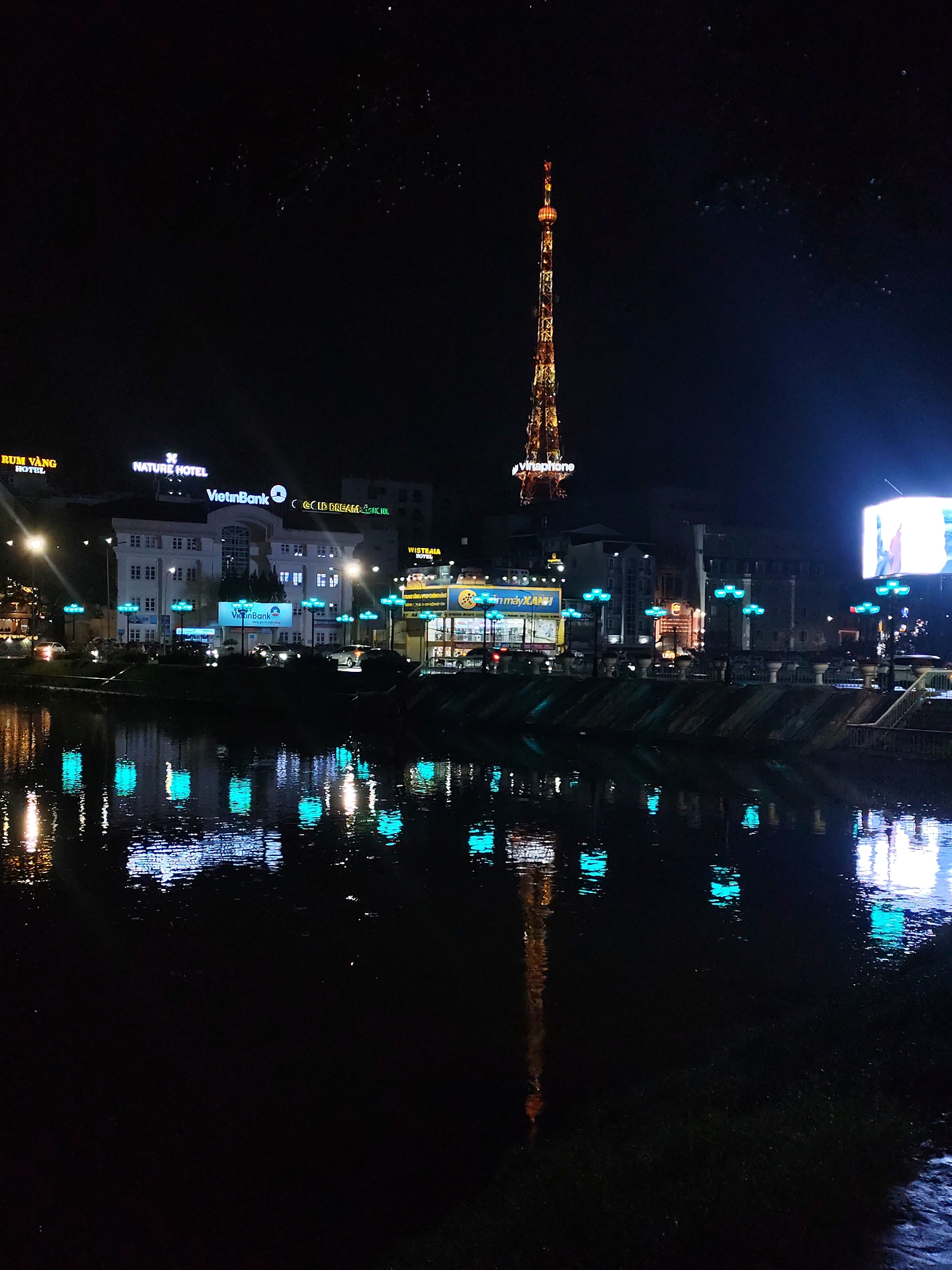 The image captures a nighttime cityscape in Da Lat, Vietnam, reflected in a dark river. Prominent features include a brightly lit telecommunications tower labeled "Vinaphone" and several buildings with neon signs advertising "Nature Hotel," "VietinBank," and "Gold Dream Hotel." Turquoise streetlights line the riverbank, and their reflections create patterns in the water. Cars and pedestrians are visible along the riverfront. The scene is illuminated by the various light sources, against a very dark sky.