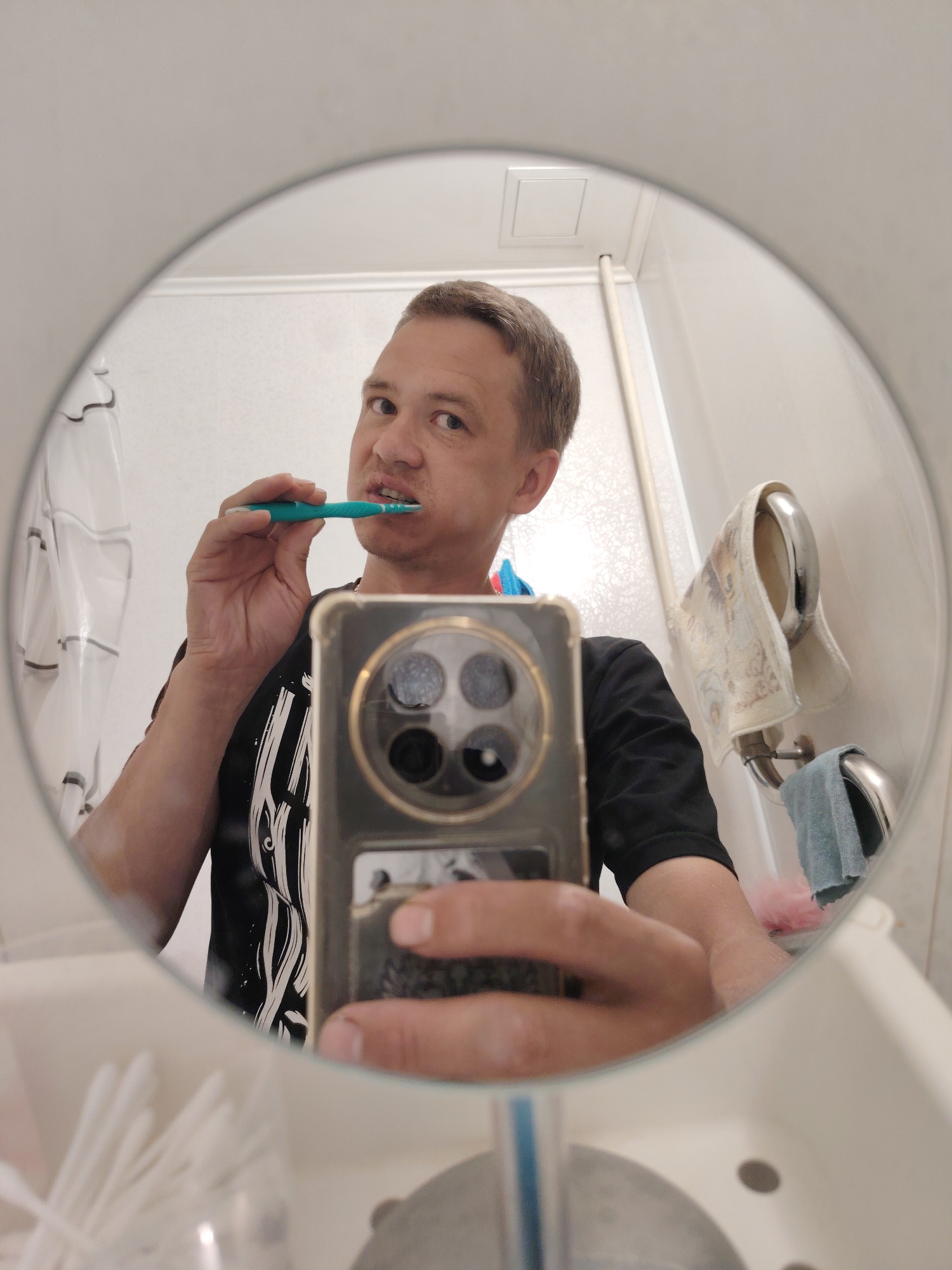 A man is brushing his teeth in a bathroom, reflected in a round mirror. He is holding a teal toothbrush in his right hand and is wearing a black t-shirt with a white graphic print. His phone, with a gold-rimmed case, is visible in his left hand, also reflected in the mirror. The bathroom appears to be a typical home bathroom, with a shower curtain visible on the left, a towel rack with a patterned towel and a blue hand towel on the right, and a sink with cotton swabs in a holder at the bottom of the frame. The lighting suggests indoor lighting, possibly from overhead fixtures. There are no discernible external environmental elements that would place this scene in a specific city or location.