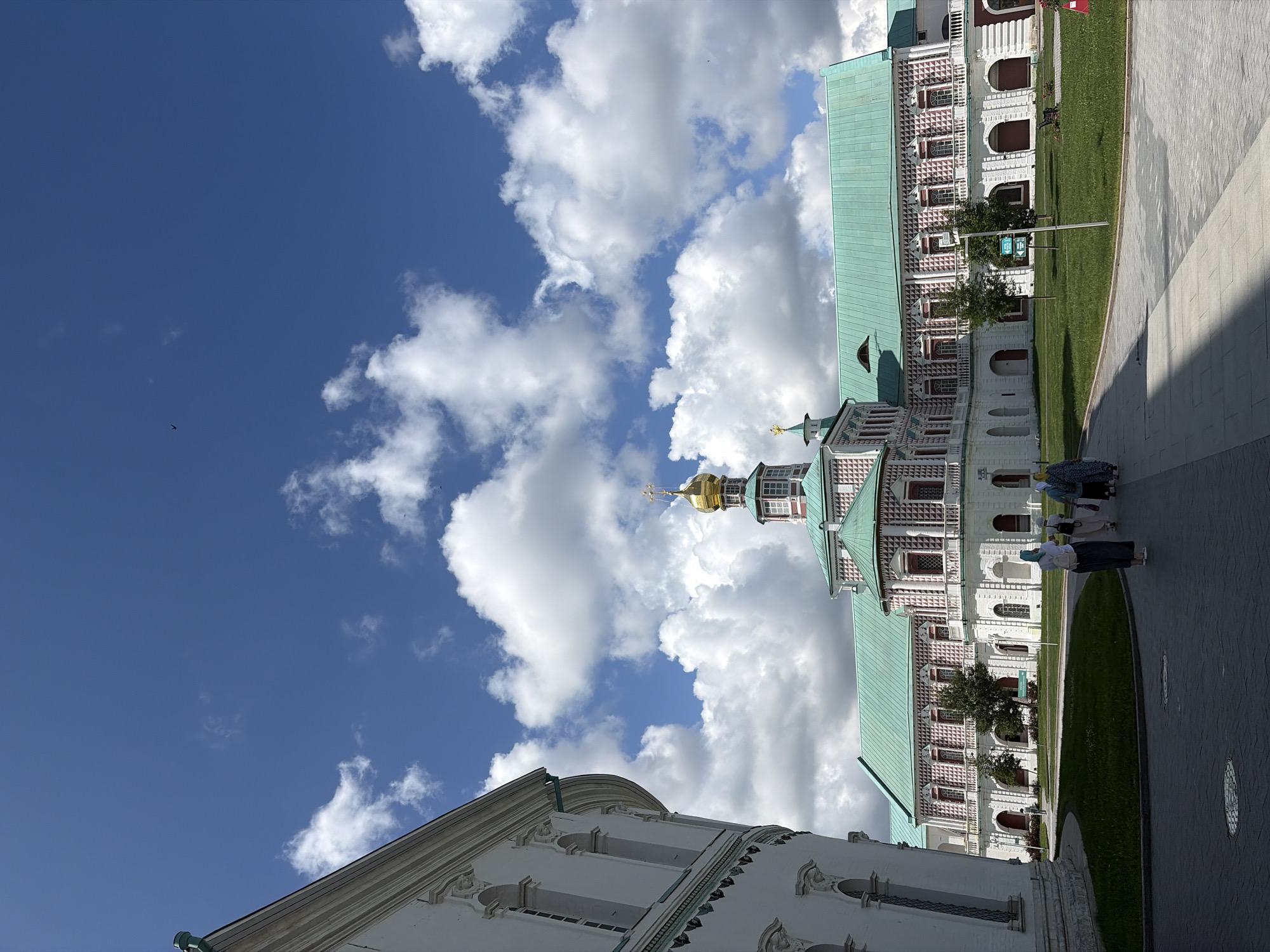 The image shows the Voskresensky New Jerusalem Monastery in Istra, Russia. The monastery complex is characterized by its white walls, red and white patterned facade, and distinctive green roofs, topped by a golden dome. The main building, likely a cathedral or church, is the focal point, with a central golden dome and smaller green spires. In the foreground, a paved walkway leads towards the monastery, with a well-maintained lawn to the right. Several people, dressed in modest attire, are walking away from the camera on the pathway. The sky is a clear blue with scattered white clouds, suggesting a pleasant day. The architecture is ornate, featuring arched windows and decorative elements.