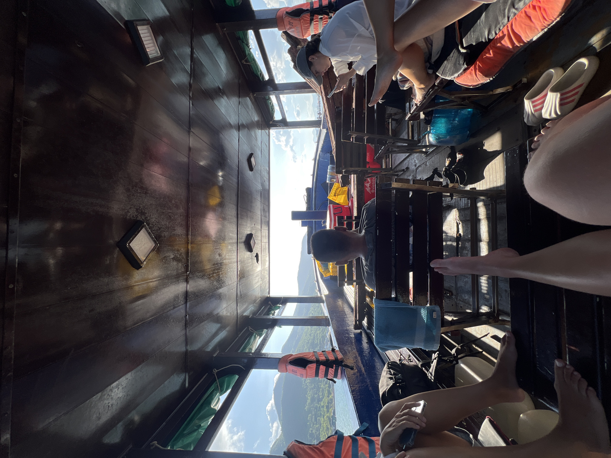 Inside a long boat with dark wood interior, various passengers are seated and visible. A man with short dark hair sits on a wooden bench. Other passengers' legs and feet are partially visible, indicating they are also seated. Life vests are attached to the boat’s frame. Through the boat's windows, a view of the water, land, and sky is visible, suggesting the boat is moving on the water. The location is the city of Nam Nha Trang Ward, Vietnam.
