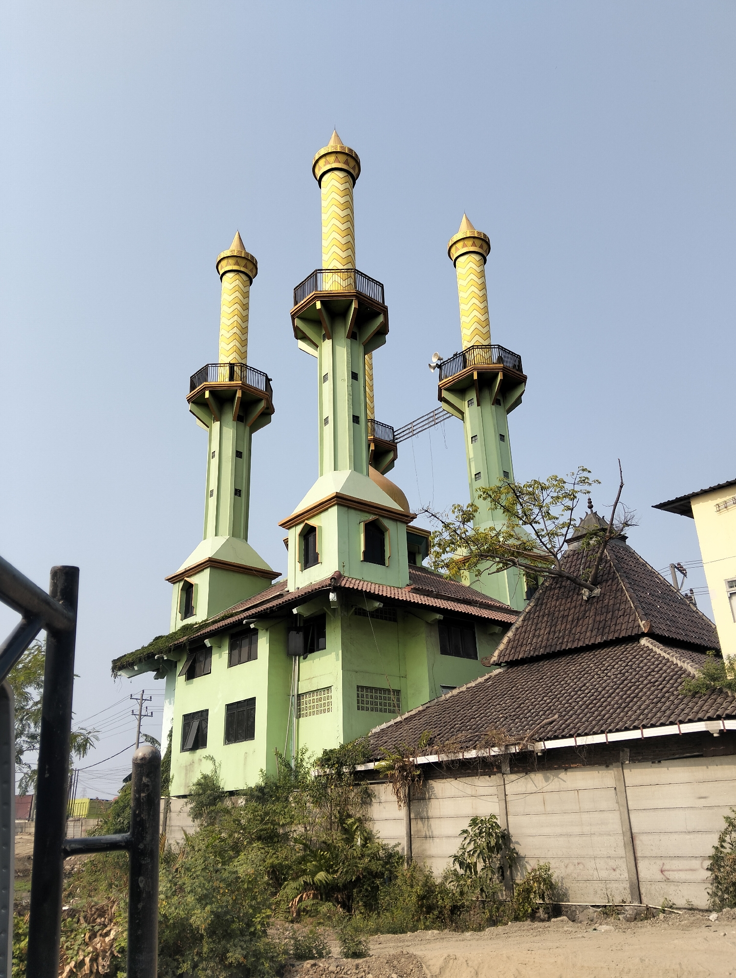 The image captures a mosque in Semarang, Indonesia, featuring a prominent green building with three tall, slender minarets. The minarets are topped with golden, patterned conical roofs and feature small balconies. The main structure of the mosque is also green, with a dark-tiled roof and multiple windows. In the foreground, a dark metal fence partially obstructs the view. To the right of the mosque, a traditional Javanese-style house with a dark-tiled hip roof is visible, along with a concrete wall and vegetation. The sky is clear and blue, suggesting daytime. No people are present in the scene.