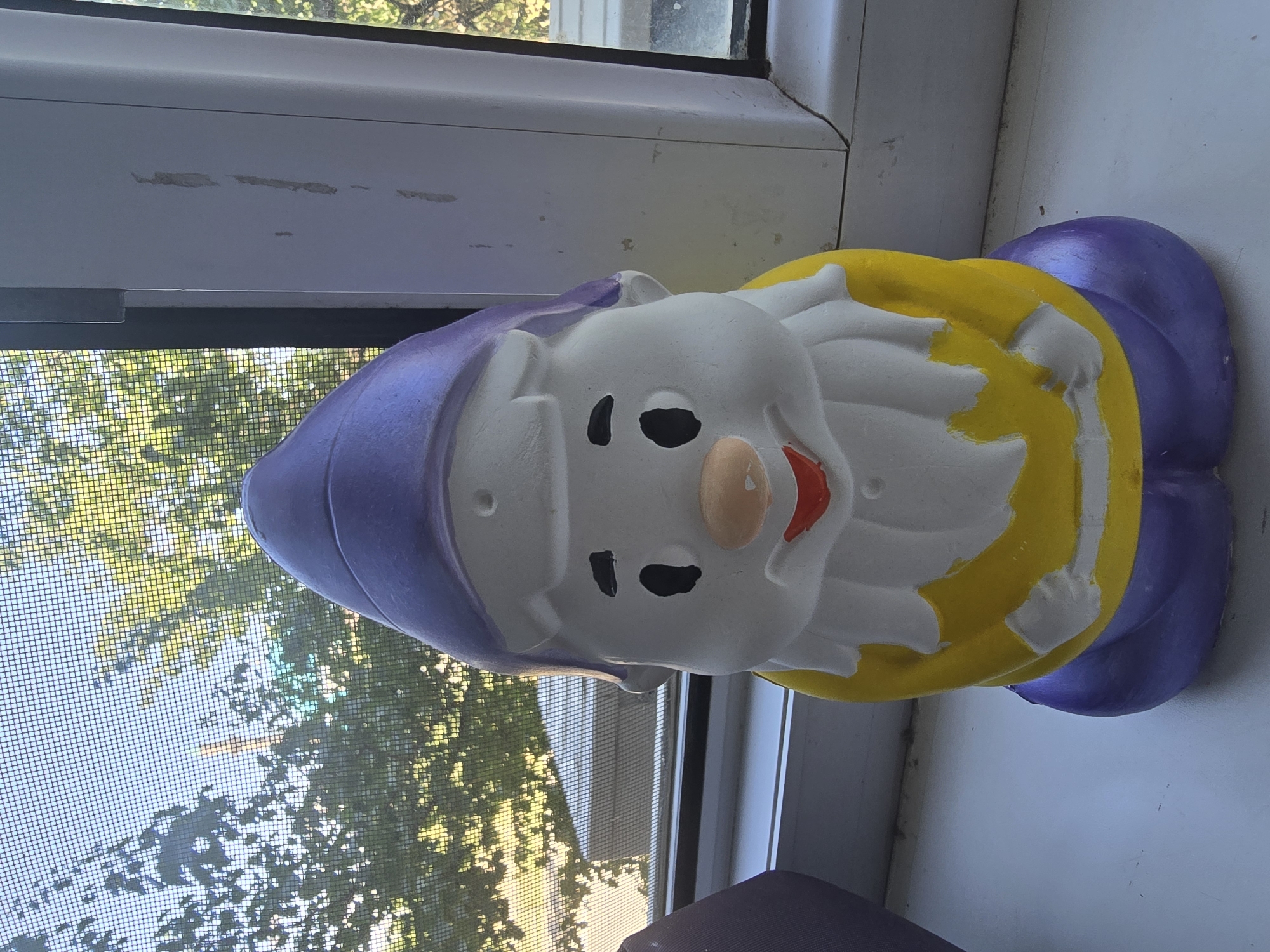 The image shows a gnome figurine positioned on a windowsill. The gnome has a purple hat, a white face with black eyes, a peach-colored nose, and a red mouth. It is wearing a yellow shirt and purple pants. The windowsill is white and appears to be part of a window with a screen through which trees and a building are visible. The scene is indoors, with the gnome facing forward. The photograph was taken on a sunny day, as indicated by the light filtering through the window and the shadows. The location of Qaratau, Kazakhstan is not visually identifiable in this image.