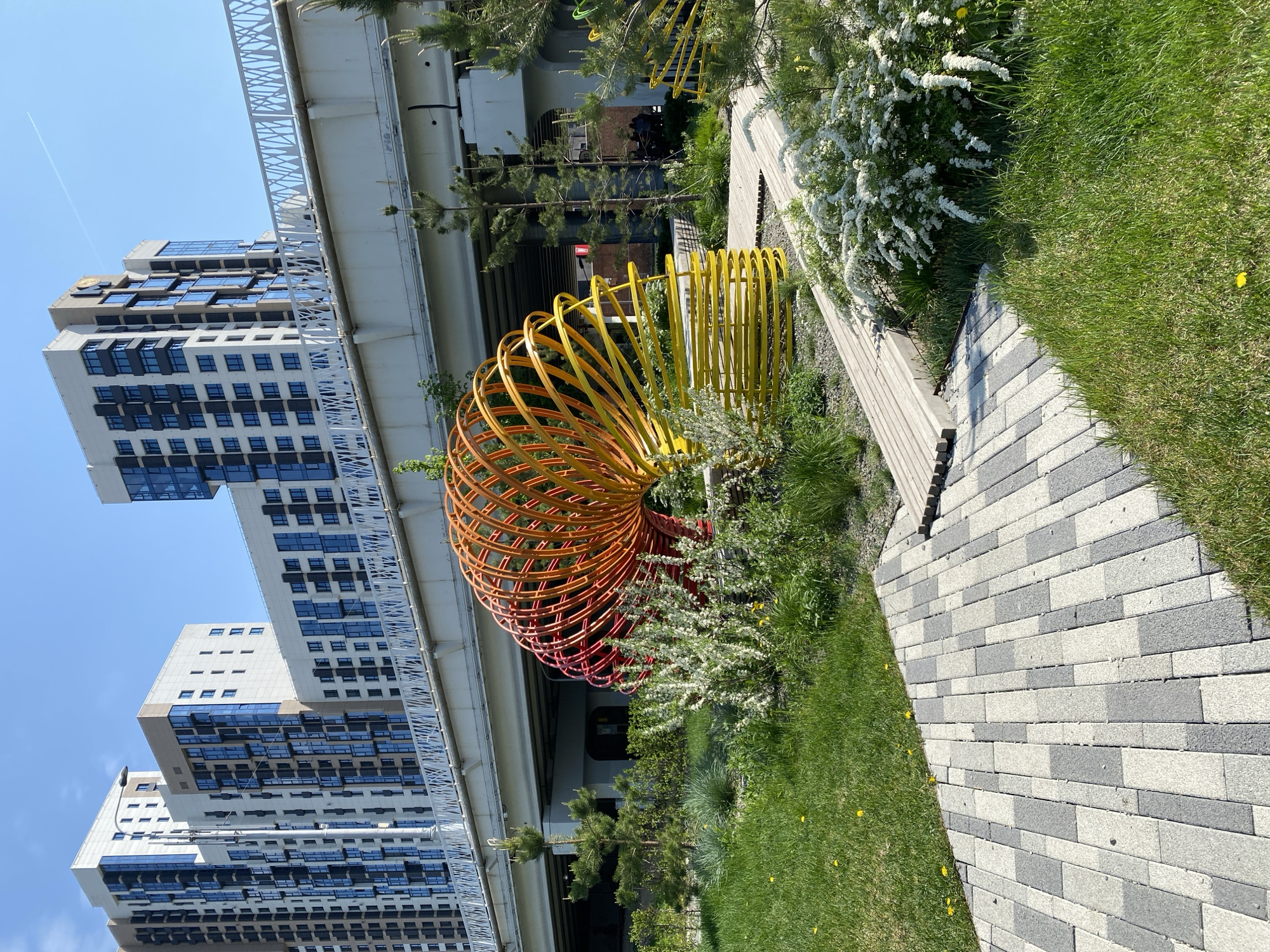 This image shows a public space in Yekaterinburg, Russia, featuring a modern architectural sculpture. The sculpture is spiral-shaped and made of painted metal, with the colors transitioning from yellow to orange to red. It is situated amidst greenery, including grass, bushes, and small trees. A paved walkway made of gray and white rectangular blocks leads alongside the sculpture.
In the background, a multistory building with a modern glass and concrete facade is visible, partially obscured by an elevated structure, possibly a bridge. The sky is clear and blue. No people are visible in the immediate foreground. The environment suggests a designed urban landscape, possibly a park or a recreational area near a residential or commercial complex.