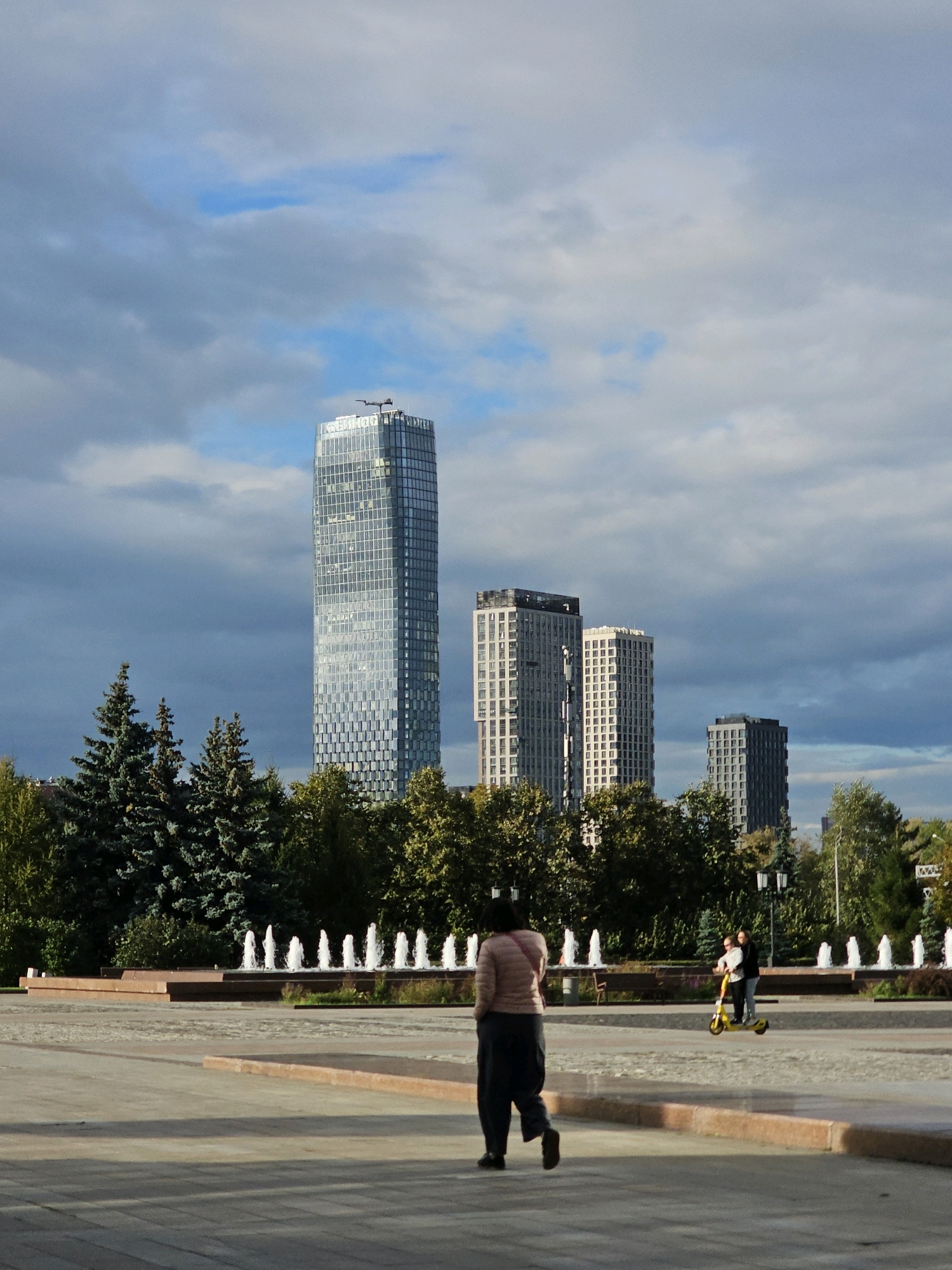 The image captures a wide view of an urban park on a cloudy day. In the background, a tall, modern skyscraper with a glass facade dominates the skyline, alongside other contemporary buildings. In the middle ground, a line of fountains is active, set against a backdrop of trees and more buildings. In the foreground, a person walks away from the camera, dressed in dark pants and a light pink jacket. To the right, two people are seen riding a yellow scooter. The park features paved walkways and raised planters, suggesting a public recreational area. The overall impression is one of a modern city park with architectural elements and active city life.