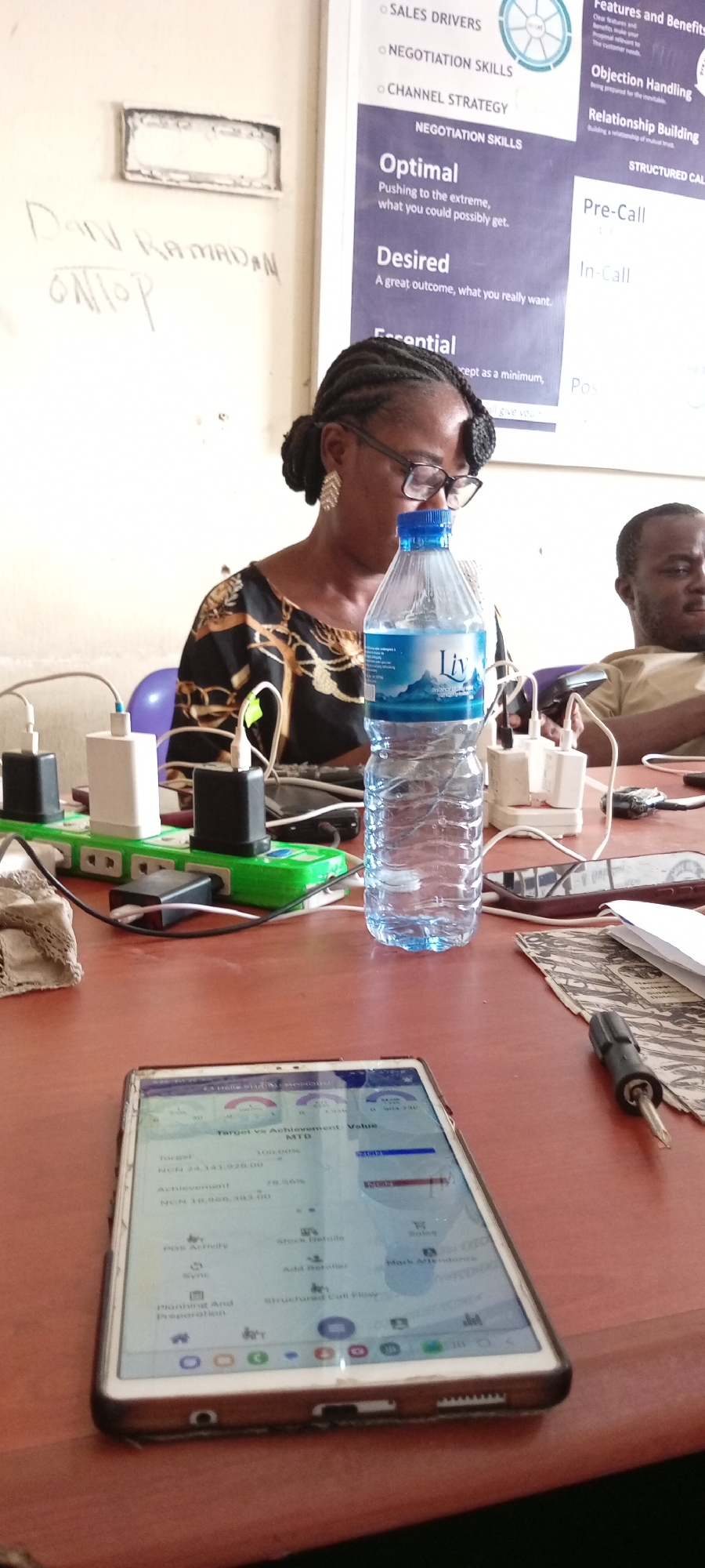 This media file captures an indoor scene with two people at a table, likely in an office setting in Fagge C, Nigeria. The primary subject in the foreground is a woman wearing glasses and a patterned black and gold dress, with her hair in braids. She is looking down at a tablet device on the table, which displays a sales-related dashboard with charts and figures like "Target vs Achievement: Value MTD," "Target," and "Achievement." Several power adapters and a green extension cord are visible, indicating charging activity. A bottle of water with a "Liv" label is placed prominently on the table.
In the background, a portion of a man's face is visible as he looks downwards, also seemingly engaged with something on the table or off-camera. Behind the subjects, a whiteboard or poster is mounted on the wall, featuring text related to sales and negotiation strategies, including sections like "SALES DRIVERS," "NEGOTIATION SKILLS," "Optimal," "Desired," "Objection Handling," and "Relationship Building." Some handwritten text is visible on the wall to the left of the poster, reading "DAN RAMADAN ONTOP."
The overall impression is a focused work or meeting environment, possibly during daytime given the ambient lighting. The presence of the sales-related materials and the tablet suggests a business context, where the individuals are likely engaged in work-related tasks.