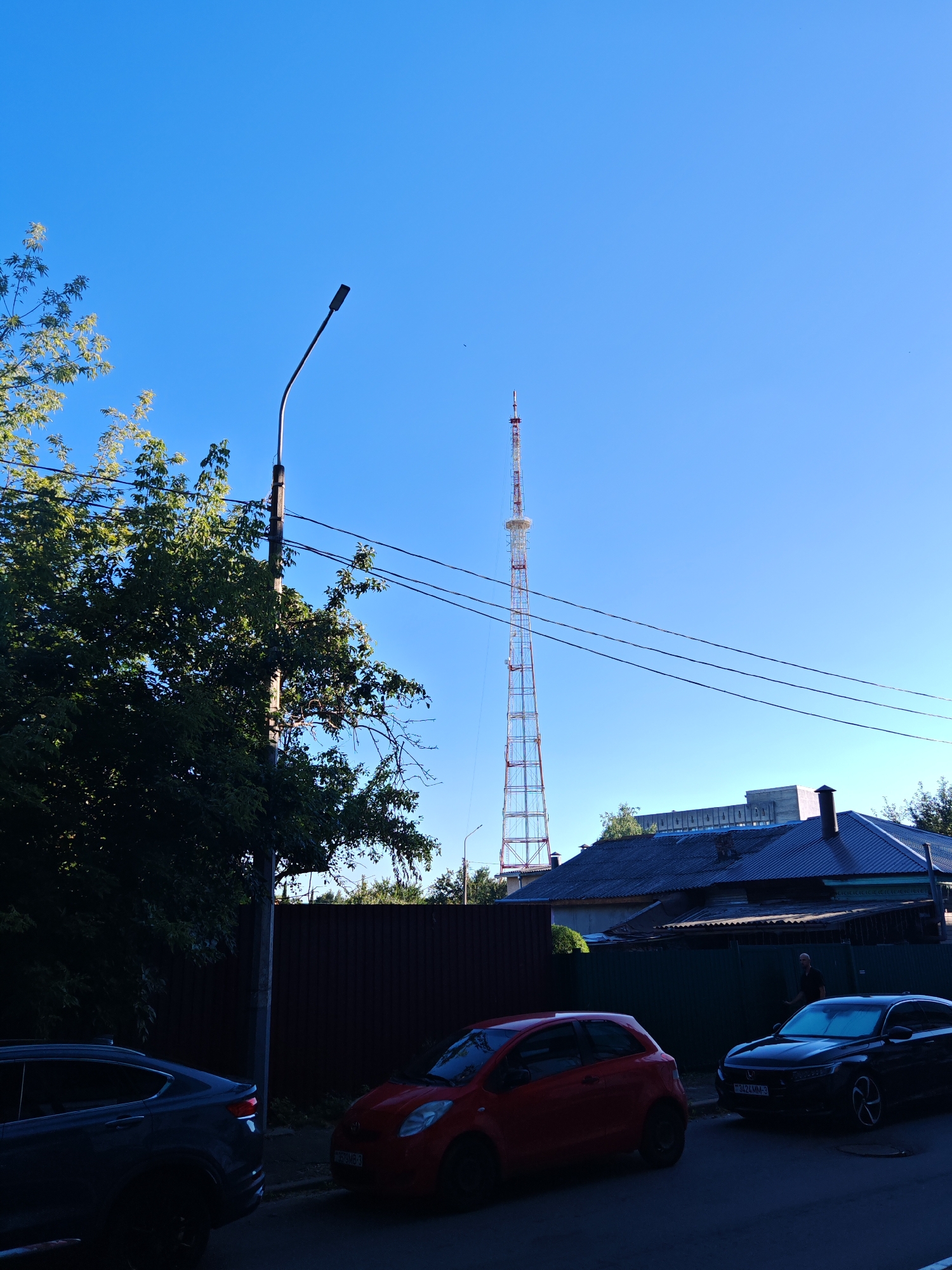 The image, taken in Homyel, Belarus, depicts a street scene under a clear blue sky. A tall, lattice-like radio tower with red and white sections dominates the background. In the middle ground, a low-rise building with a dark, sloped roof is visible, partially obscured by trees and a dark brown fence. A red hatchback is parked on the street in the foreground, to the left of a dark-colored sedan. A person is visible standing near the dark sedan. Several power lines are strung across the sky, supported by a lamppost on the left side of the frame. Lush green foliage from trees borders the left and right sides of the image, with shadows indicating the sun is relatively high in the sky.