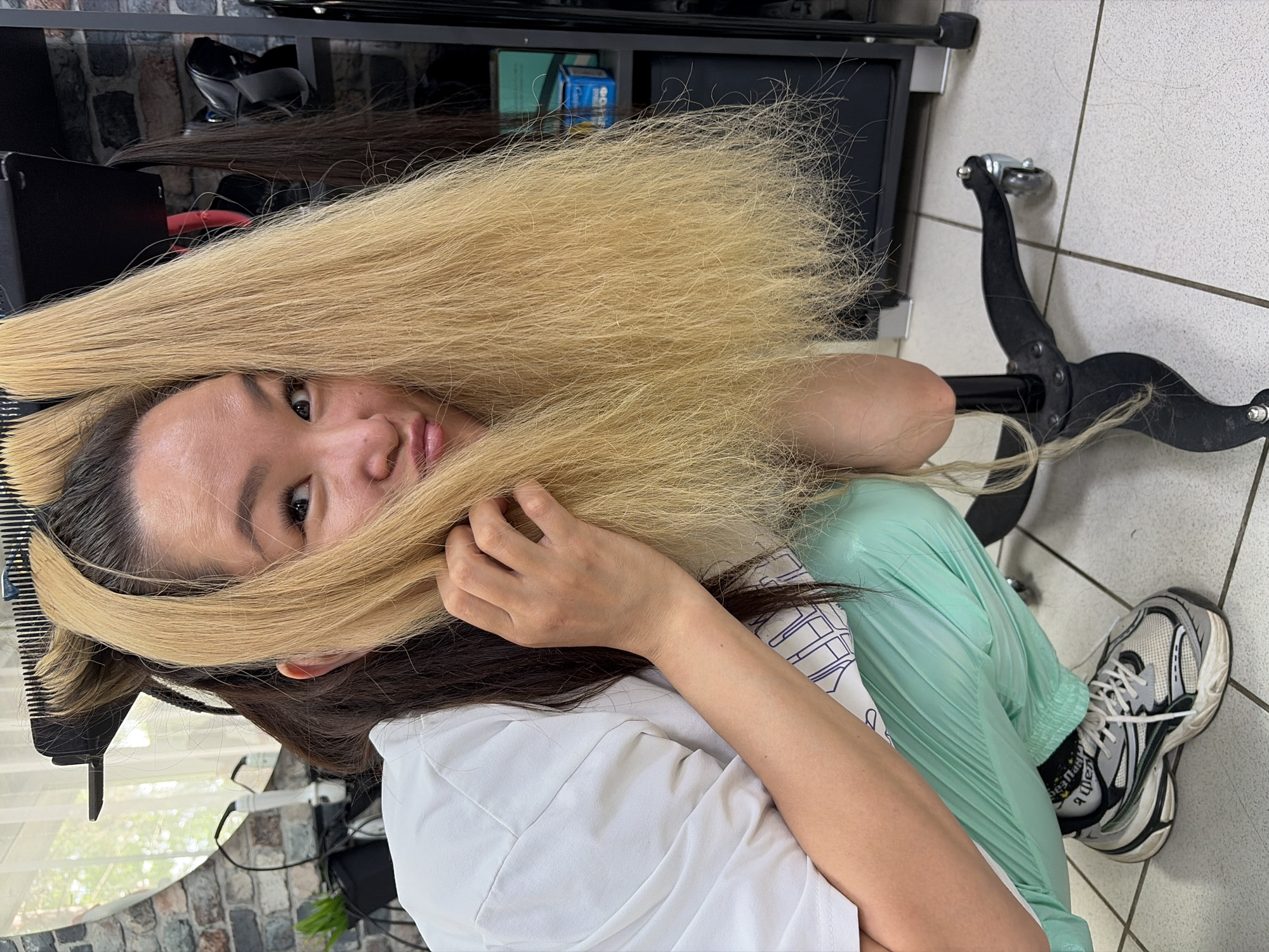 A young Asian woman with dark brown hair and blonde extensions is squatting on a tiled floor. She is wearing a white t-shirt, light green sweatpants, and athletic shoes. Her right hand is touching her hair, which is pulled forward over her face. She is looking at the camera with a playful expression. The background shows a hair salon with shelves, styling equipment, and a mirror. The image appears to be taken indoors, possibly in Chaikovsky, Russia.