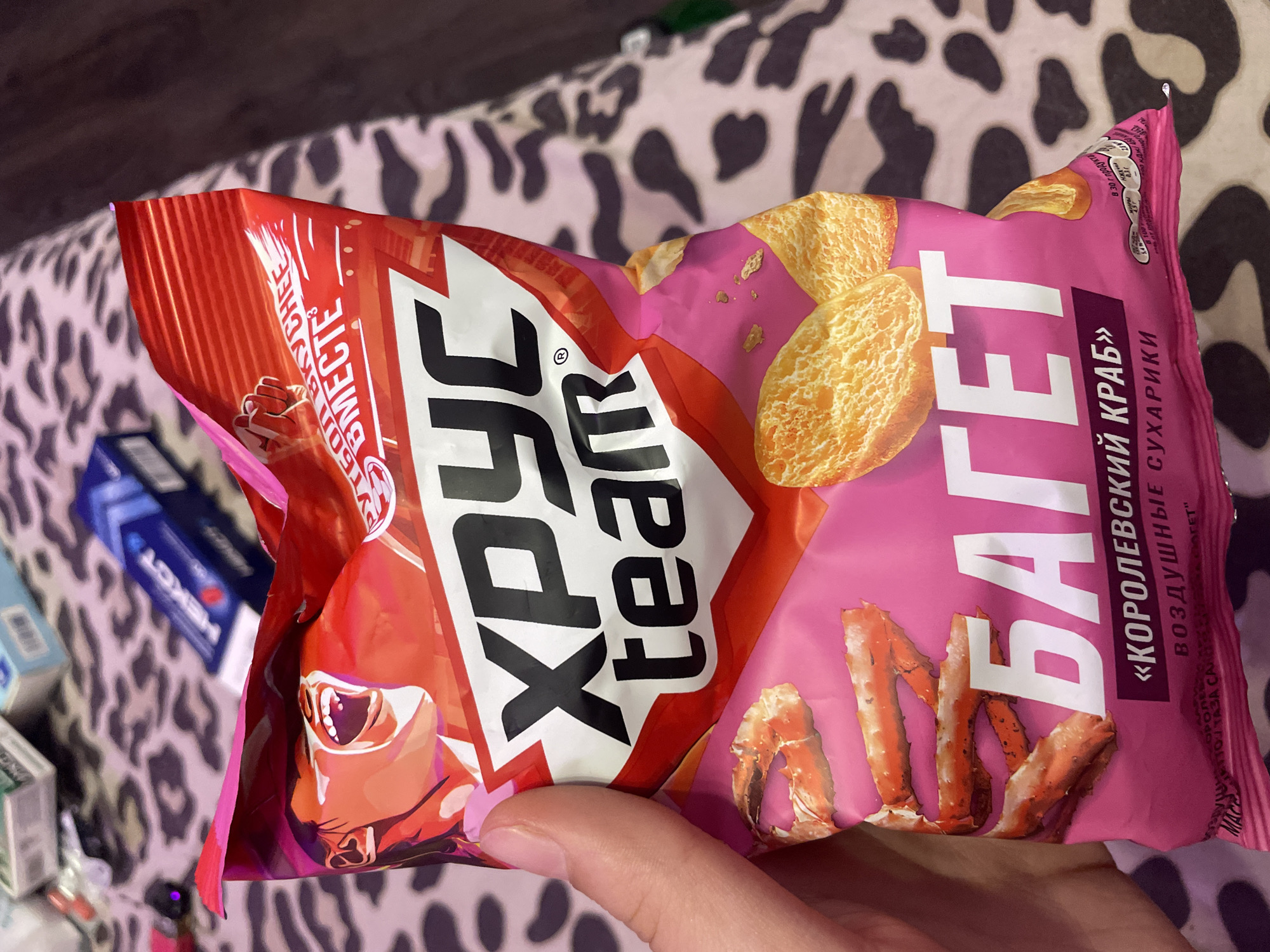 A hand is holding a brightly colored snack bag featuring "ХРУСТ team" and "БАГЕТ" in large white lettering against a pink and red background. Images of crab legs and toasted bread slices are also visible. The background appears to be a patterned fabric, possibly a blanket or bedding, with a blurred assortment of items behind it, including what appear to be cigarette packs and a blue tube. The image does not contain any identifiable landmarks or contextual clues to suggest a specific location within the city of Vaganovo, Russia.