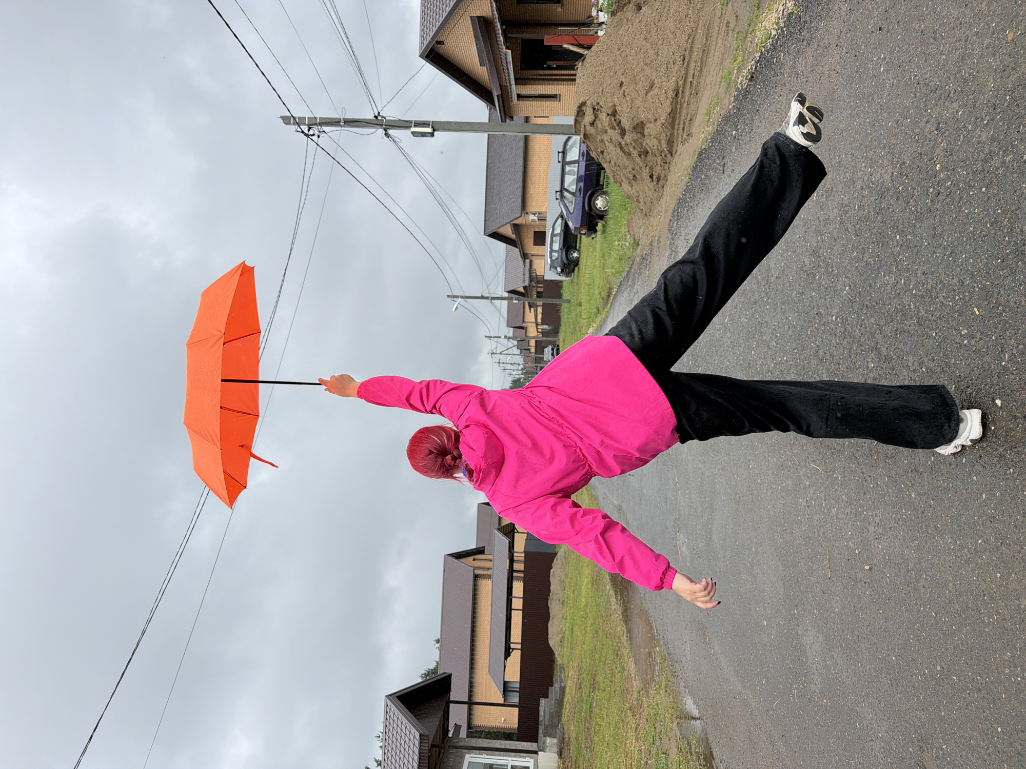 A person with bright red hair, wearing a pink jacket and black pants, stands on a wet asphalt road holding an open orange umbrella above their head. Their right arm is extended upwards, holding the umbrella's handle, while their left arm is outstretched to the side. Their left leg is bent and lifted off the ground, with the right leg extended backward. The background shows a row of houses with brown tiled roofs, some under construction, on a cloudy day. Several cars are parked along the road, and utility poles with overhead wires are visible. A pile of sand sits to the right of the road. This scene is located in a residential area of Chaikovsky, Russia.