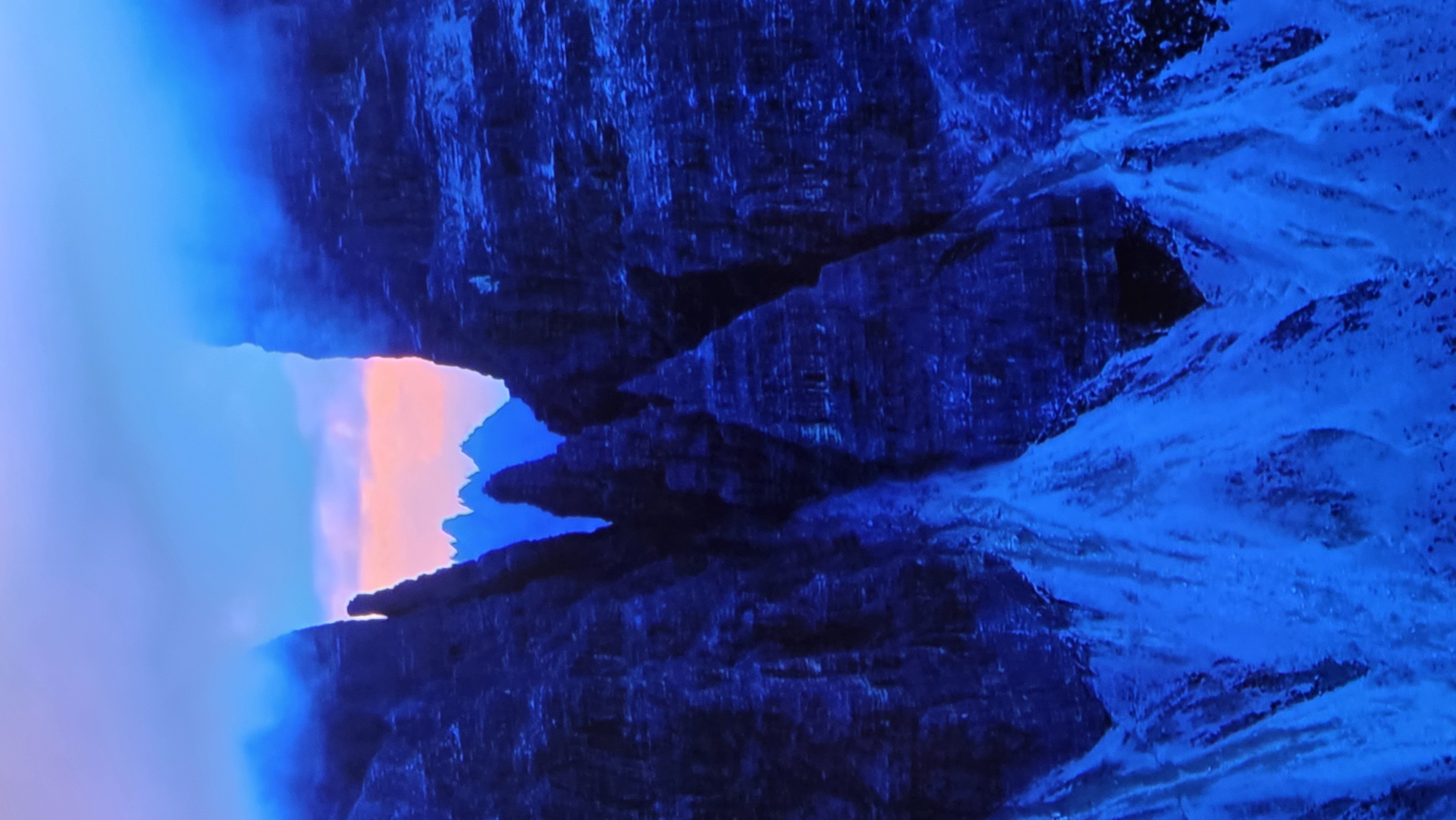 The image depicts a dramatic mountain landscape at dusk or dawn. The foreground is dominated by dark, jagged rock formations rendered in deep blues and purples, with streaks of bright blue, possibly ice or snow, cascading down the slopes. These formations create a sense of depth and a narrow passage or canyon. In the background, the sky is a gradient of soft blues and pinks, suggesting a twilight atmosphere. Several sharp, pointed peaks rise in the distance, silhouetted against the colorful sky. The overall impression is one of rugged, imposing natural beauty. This image does not contain any identifiable features or context that would place it in the city of Batumi, Georgia.