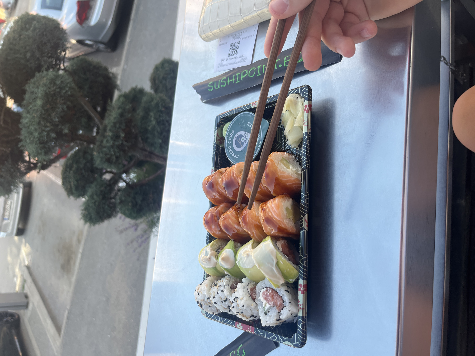 A hand is shown using chopsticks to pick up a piece of sushi from a takeout container. The sushi is arranged in three rows within a black plastic tray. The top row appears to be salmon rolls glazed with a dark sauce. The middle row contains rolls with avocado and what looks like a creamy sauce. The bottom row consists of rolls covered in black sesame seeds, with one piece showing a pink filling. There is also a small container of pickled ginger and a small green packet with a white lid, possibly soy sauce or wasabi, in the tray. A wooden chopstick rest is also visible.
In the background, a black sign with green lettering reads "SUSHIPOINT.BG". Below this sign is a small receipt with a QR code and some text in Cyrillic script. To the left of the sushi tray, there are decorative artificial evergreen topiaries. The surface the sushi is placed on appears to be a brushed metal countertop. In the far background, a silver car is partially visible. The lighting suggests it is daytime, possibly outdoors given the greenery and car, with the sun casting some reflections on the metallic surface. The overall scene suggests a casual meal or snack being prepared for consumption. The location context provided is Sofia, Bulgaria.