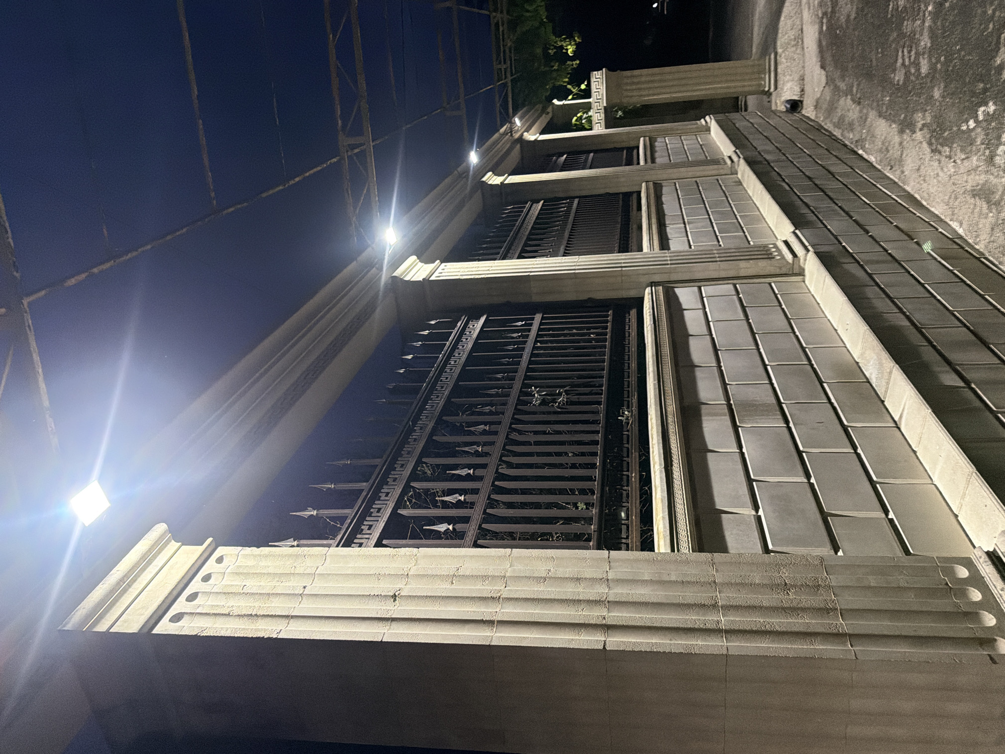 The image captures a nighttime view of a stone wall and an ornate metal fence, illuminated by artificial lights. The wall, constructed from large, light-colored rectangular blocks, is topped by a decorative ledge. A dark metal fence with pointed finials and intricate patterns runs along the top of the wall. Several classical-style stone columns, fluted and with elaborate capitals, support an overhead structure. The sky above is dark blue, with visible supports for a canopy or roof. The ground in the foreground is paved and appears wet in places. The scene suggests an entrance or boundary of a well-maintained property, possibly a historic or upscale building. The lighting creates strong highlights and shadows, emphasizing the architectural details. There are no people or animals visible in the image. The context of Sochi, Russia, implies a potentially coastal or resort setting, which might influence the architectural style.