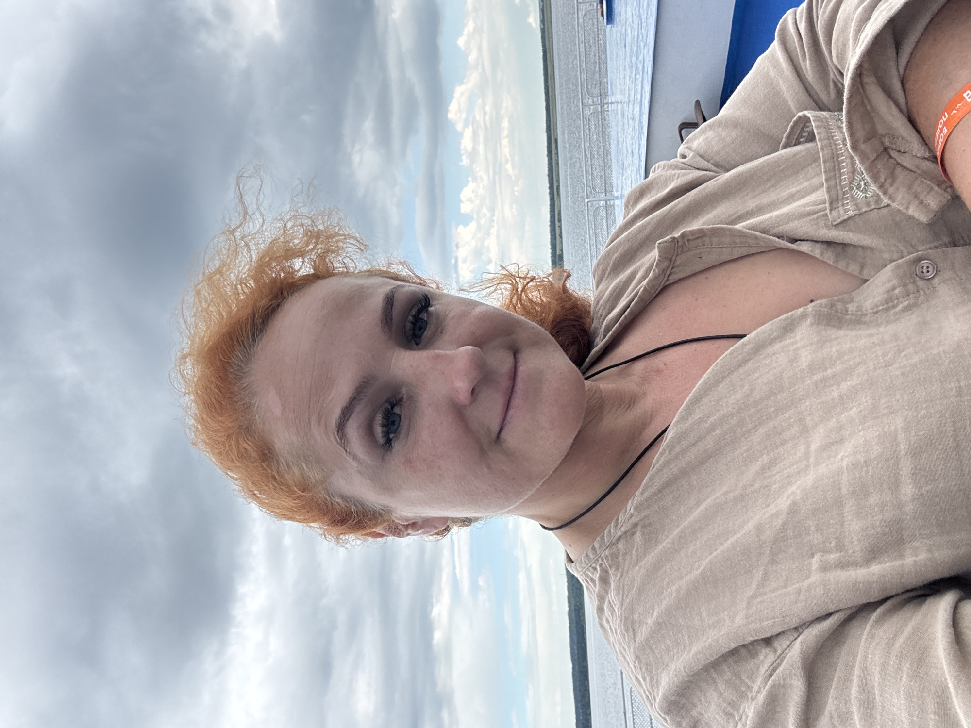 A woman with red curly hair and fair skin, wearing a tan button-up shirt, takes a selfie on a boat. She is smiling slightly and looking at the camera. Her hair is windswept. She is wearing a black cord necklace. In the background, a body of water is visible with a treeline on the horizon under a cloudy sky. The foreground shows the railing of the boat and a blue surface, possibly water. The location is Ikskoe Ust'e, Russia.