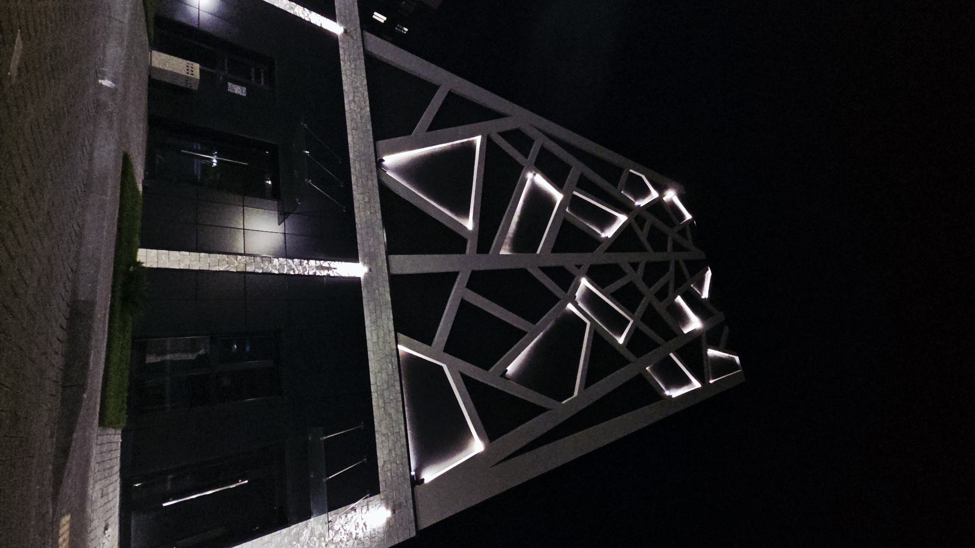 This is a low-angle, outdoor, nighttime photograph of a modern building with distinctive geometric architectural lighting. The building's facade is a dark color, possibly black, with large square or rectangular panels. Above this dark section, the building has a white, geometric lattice-like structure. Within the spaces of this white structure, white lights are embedded, creating illuminated geometric shapes against the dark night sky. The lighting is concentrated and appears to be recessed.
The lower portion of the building features large glass doors and windows. Two prominent vertical light fixtures, one on each side of the central entrance area, illuminate the stone-like texture of the building's exterior walls. The ground in front of the building is paved with cobblestones, and there is a strip of green grass adjacent to the pavement. The sky is completely dark, indicating nighttime.
The building is located in the city of Ulyanovsk, Russia. There are no people visible in the photograph. The image captures the modern design and striking nighttime illumination of the building.