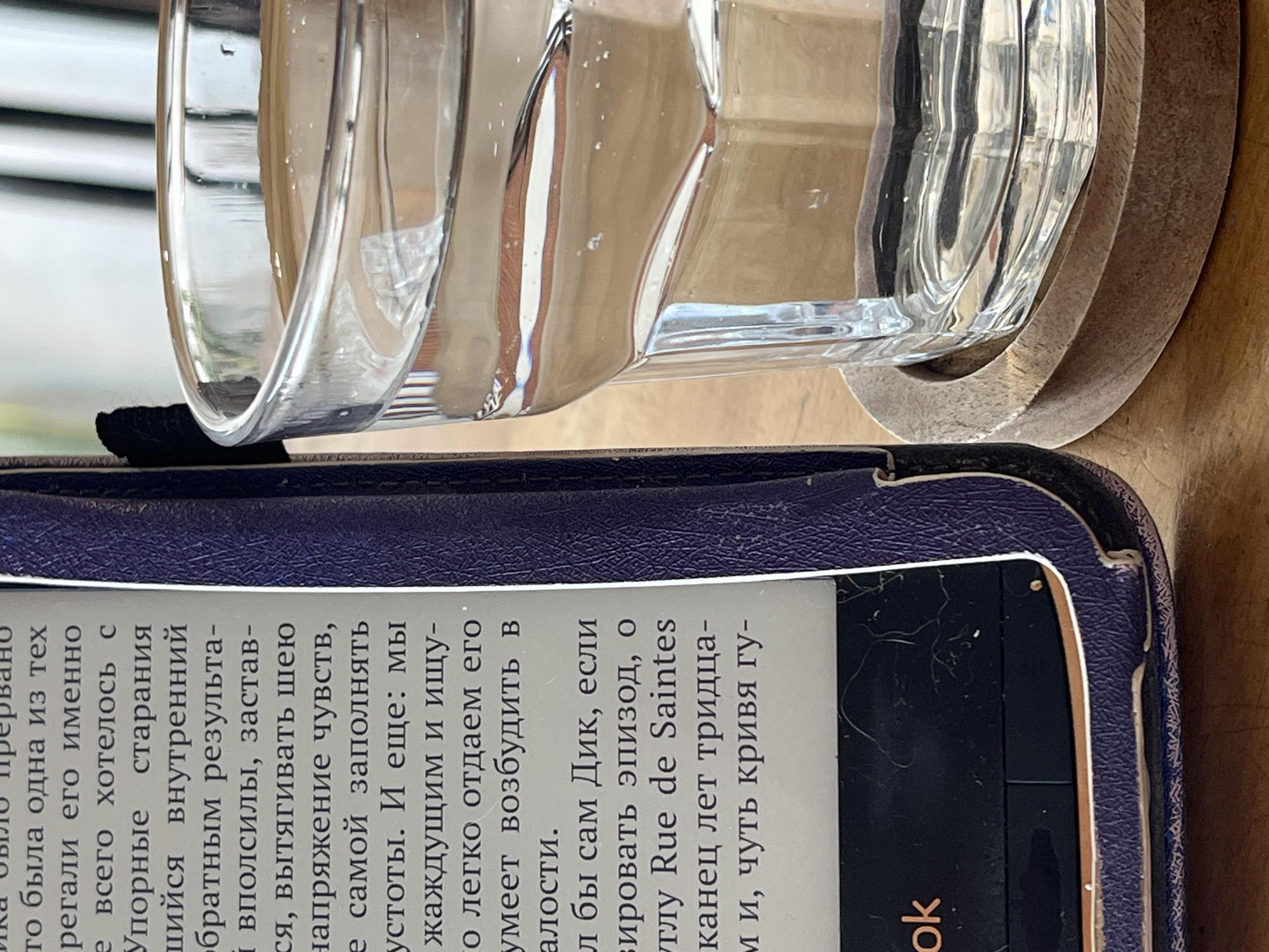 The image shows a close-up shot of an e-reader with a partially visible screen displaying Cyrillic text. The e-reader has a dark purple cover. Resting partially on top of the e-reader is a clear glass, possibly a drinking glass, sitting on a round wooden coaster. The background is slightly blurred, suggesting a close focal point on the e-reader and glass. There is no information about the location.
