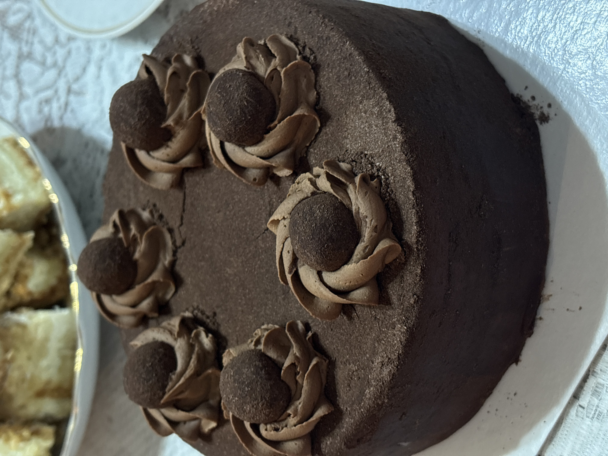 The image shows a close-up view of a round chocolate cake decorated with piped chocolate frosting rosettes, each topped with a dark chocolate truffle. The cake is placed on a white plate. To the left of the cake, a partial view of a plate containing what appears to be pastries or small cakes is visible. The background is a textured white surface, possibly a tablecloth. There is no information about this image being taken in the city of Krasnoyarsk, Russia. There are no people visible in the image.