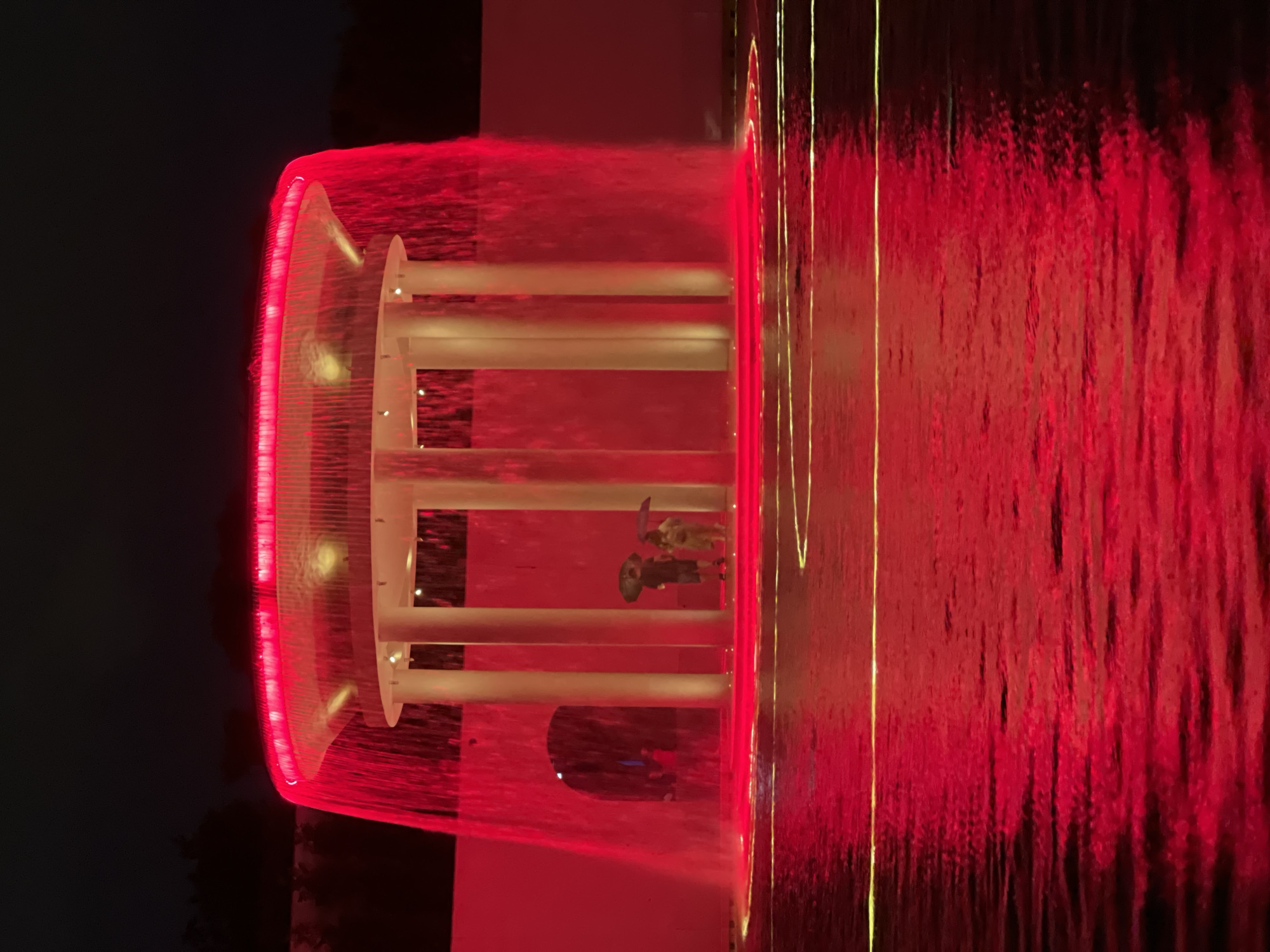 The image depicts an illuminated fountain at night. The water cascading from the fountain is a vibrant red, and the surrounding pool reflects this intense color, with faint yellow lines of light visible on its surface. A circular structure with white columns supports the top of the fountain, which is also lit with red lights. Two people, one holding an umbrella, are standing near the base of the fountain, partially obscured by the falling water. The background is dark, with a hint of trees and a curved wall. The scene is a public space, likely an outdoor plaza or park.
This image is not located in Krasnodar, Russia. It is a photograph of the "Watering the Flag" fountain in front of the Incheon National University in Incheon, South Korea.