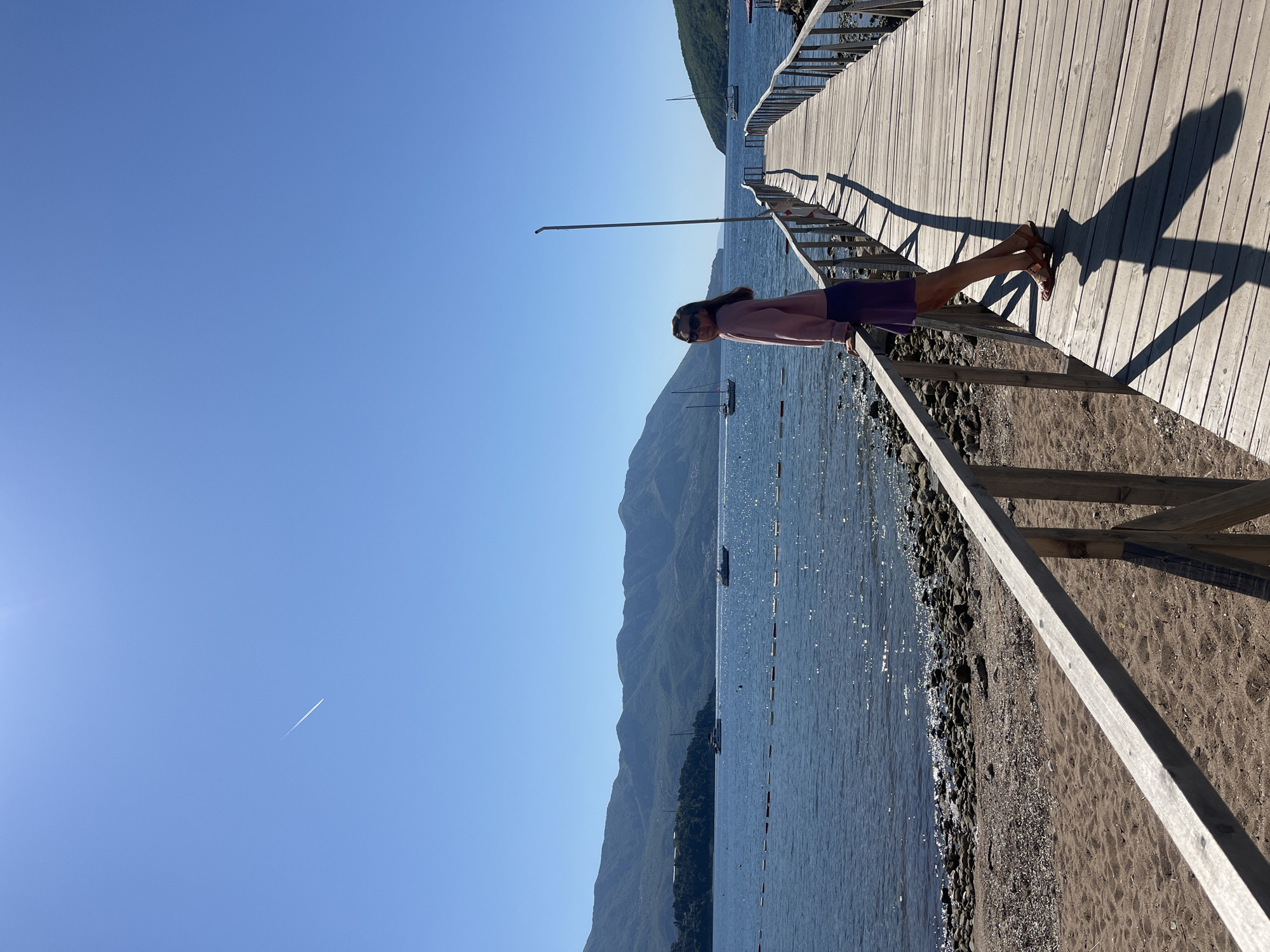 A woman stands on a pier, leaning against the railing, gazing towards a mountainous landscape across a body of water. She is wearing a light pink long-sleeved shirt, a purple skirt, and brown sandals. The pier is made of wood and has a simple wooden railing. The water is calm, and several boats are visible in the distance. The sky is a clear blue, with a contrail from an aircraft visible in the upper left corner. The scene appears to be a sunny day at a seaside location. The location is the city of Budva, Montenegro.
