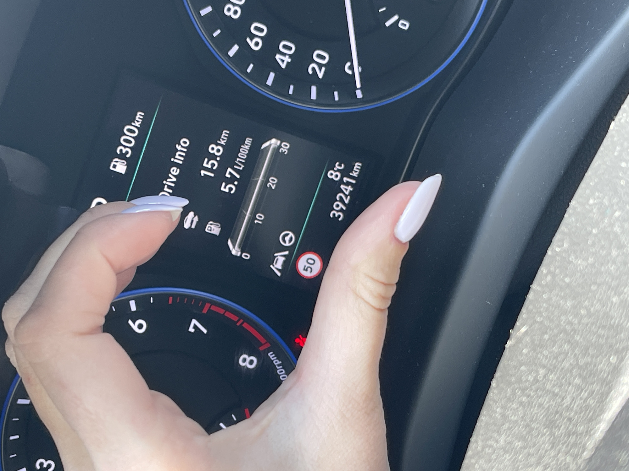 The image shows a close-up of a car dashboard with a hand partially obscuring the display. The dashboard features a speedometer with the needle pointing near 0, indicating the car is likely stationary or moving very slowly. A digital display shows various data including:
- Remaining fuel range: 300km
- Trip distance: 15.8 km
- Fuel consumption: 5.7L/100km
- Outside temperature: 8°C
- Odometer reading: 39241 km
A speed limit sign (50) is visible. The car's RPM gauge is visible. A seatbelt light is on.
Given the numerical and text data on the dashboard, along with the language being similar to Bulgarian, the image is likely taken in a location where the metric system is used, consistent with Sofia, Bulgaria.