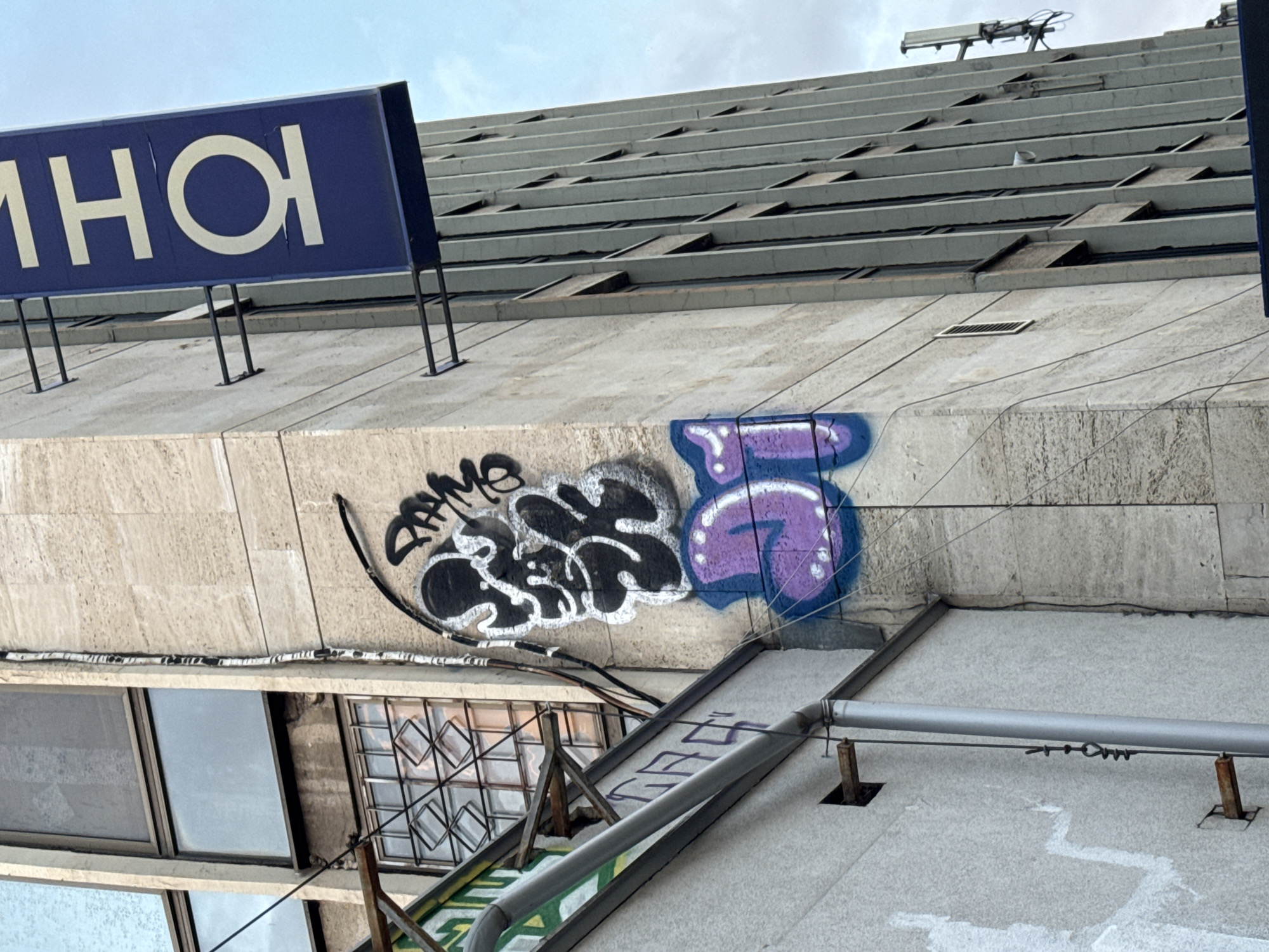 The image shows a section of a building in Sofia, Bulgaria. The building has a beige facade with visible graffiti. A blue sign with white Cyrillic letters is mounted on top. The building’s architecture features horizontal lines, possibly indicating stacked stories. There are windows and metal fixtures visible, along with rooftop equipment. The sky is overcast.