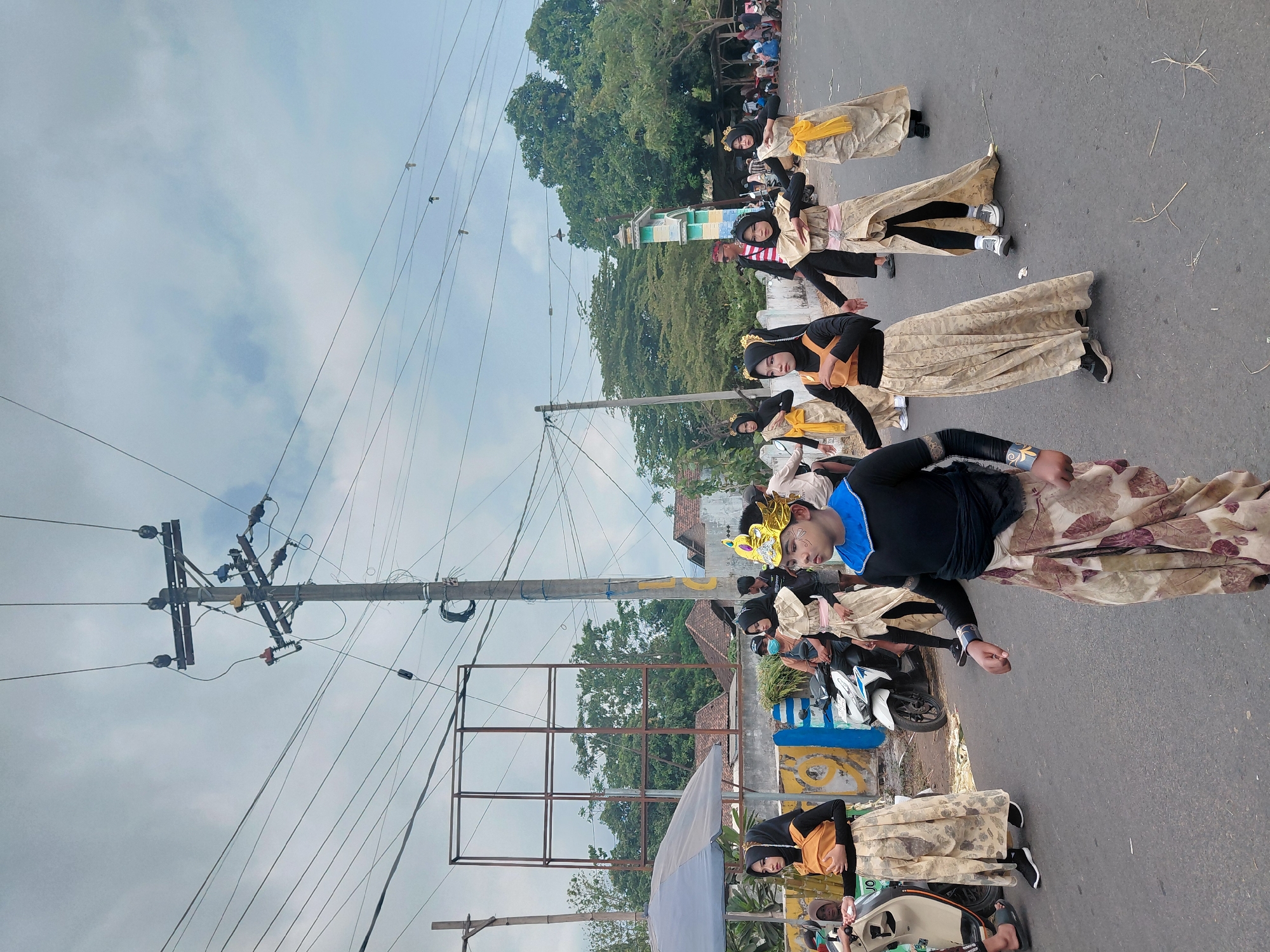 In the city of Bimo, Indonesia, a group of children and young people are participating in a parade on a paved road. The individuals are dressed in elaborate costumes, some featuring golden headdresses and flowing, patterned skirts. Some are wearing headscarves. Several are performing coordinated dance moves. They are walking on the right side of the road, with a utility pole and power lines dominating the background. Other people, some sitting or standing, are visible on the side of the road, observing the procession. Motorcycles are parked on the side of the road, and a large, empty billboard frame stands out against the trees and buildings in the distance. The sky is overcast with scattered clouds.