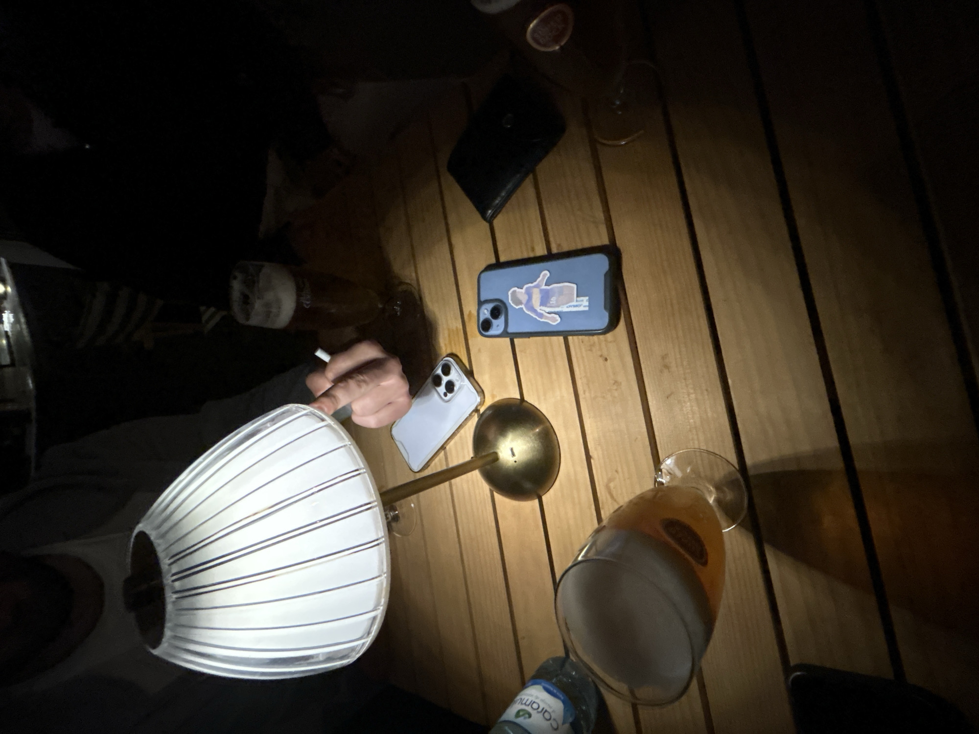 An overhead shot depicts a partially illuminated table, likely made of wood, with several items arranged on it. The scene appears to be in low light, with a lamp on the left side casting the primary light source. The lamp features a white, vertically striped shade and a brass-colored base.
Several mobile phones are visible: one with a light blue case adorned with a figure sticker, and another one with a metallic back. Next to the phones, a black wallet rests on the table.
Two beer glasses, both filled with a yellow-colored liquid, are scattered across the table. One beer glass appears to be larger than the other. A clear plastic bottle is near the bottom right corner.
A person can be seen holding a cigarette between their fingers. Based on the beer and the casual arrangement of items, the setting appears to be informal, possibly a bar or a social gathering. The location of Mirandela, Portugal is not discernible from the visual elements.
