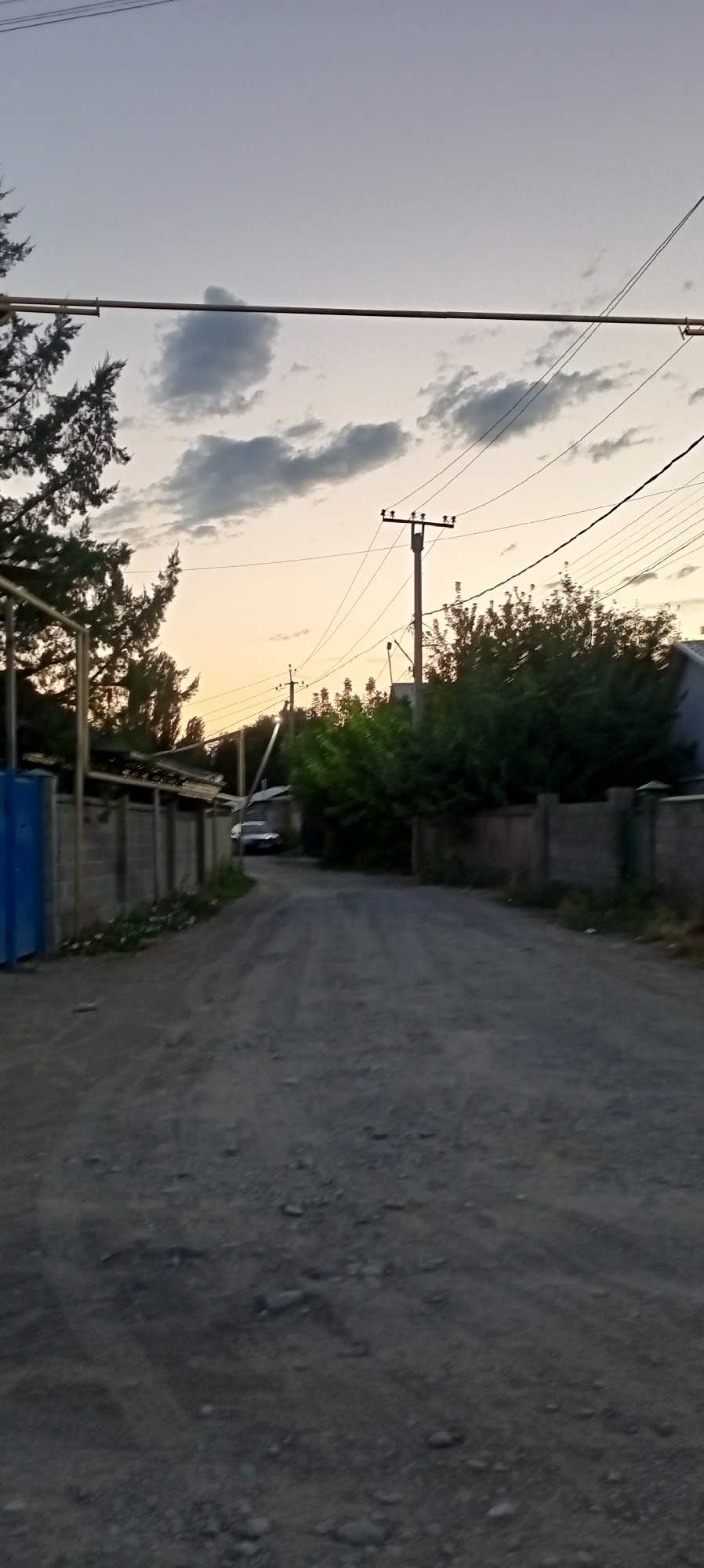The image depicts a dusty, unpaved road in the city of Romanovka, Kyrgyzstan, under a twilight sky. The road is lined with low, concrete block walls on either side. On the left, a section of a blue metal fence or structure is visible. Trees and dense green foliage line the right side of the road, partially obscuring buildings. Power lines stretch across the frame, supported by utility poles, one of which is prominently visible with multiple crossbars. A single, dark-colored car is parked or moving slowly in the distance on the road. The overall scene suggests a rural or semi-urban setting.
