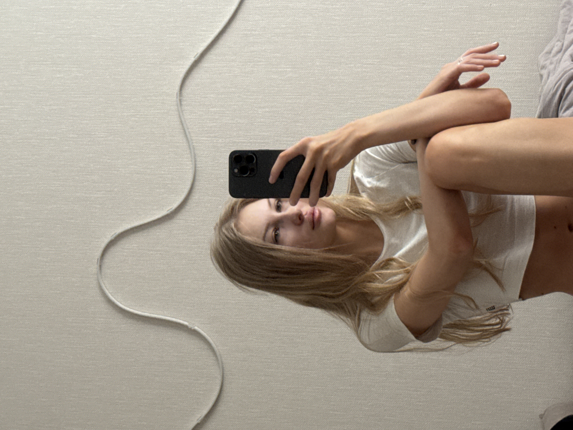 A young Caucasian woman with long blonde hair takes a selfie in a room. She is wearing a white t-shirt and is sitting with her legs crossed. She is holding a black smartphone in her right hand and is looking at the phone's screen. The background is a textured wall with a decorative wave pattern. There is no identifiable information to suggest the location is in the city of Perm, Russia.