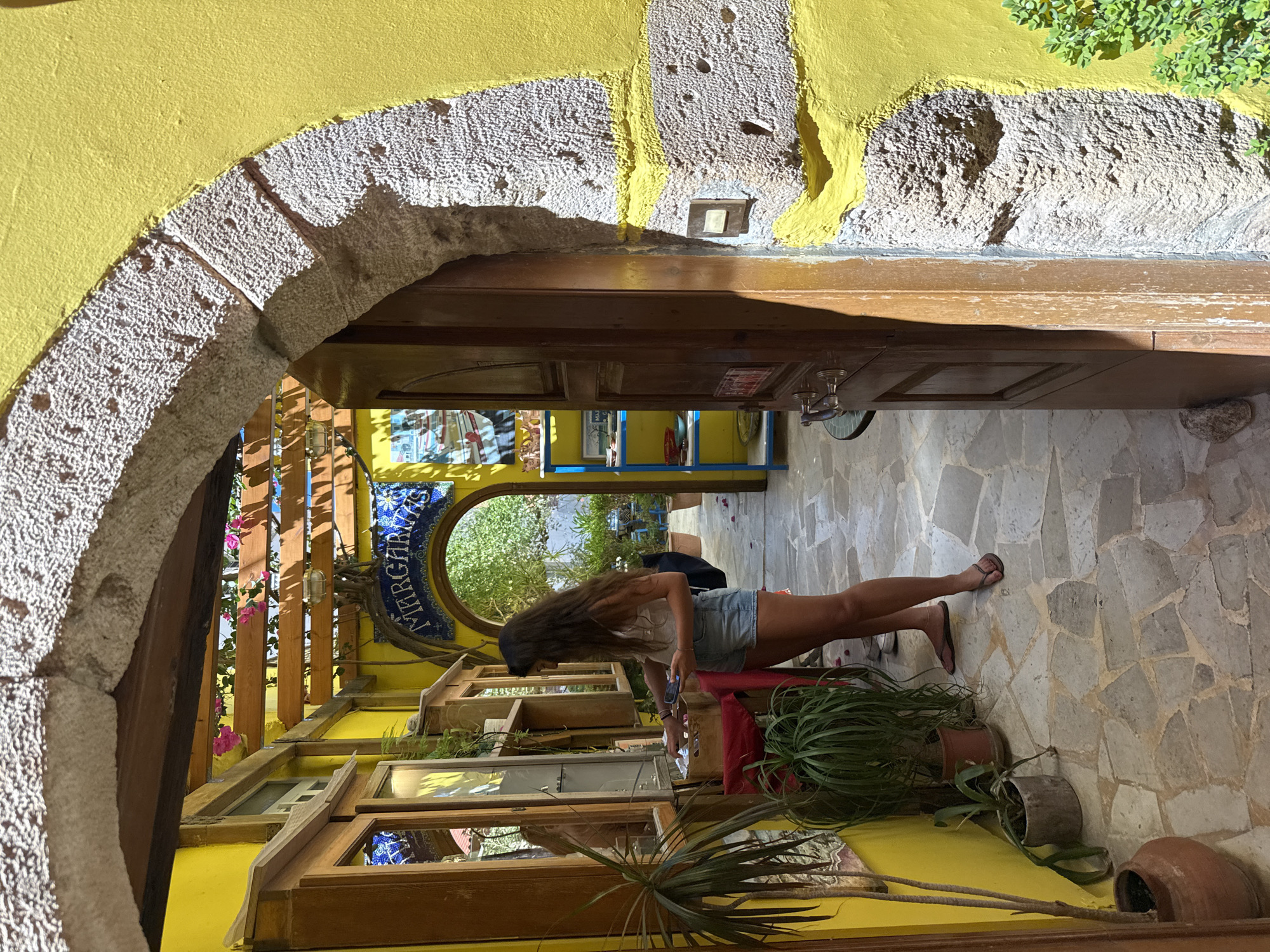 In the city of Chania, Greece, a woman with long brown hair, wearing a white top and denim shorts, stands near a wooden display shelf. She is looking down and holding a mobile phone in her left hand. The environment is a narrow outdoor walkway with brightly painted yellow walls and a stone archway. The walkway is paved with irregular stones, and several potted plants, including a large dracaena, are placed along the walls. Above, a wooden pergola with pink flowers provides shade. A mosaic sign arching over a doorway reads "MARGARITAS." The scene appears to be the entrance to a shop or restaurant.