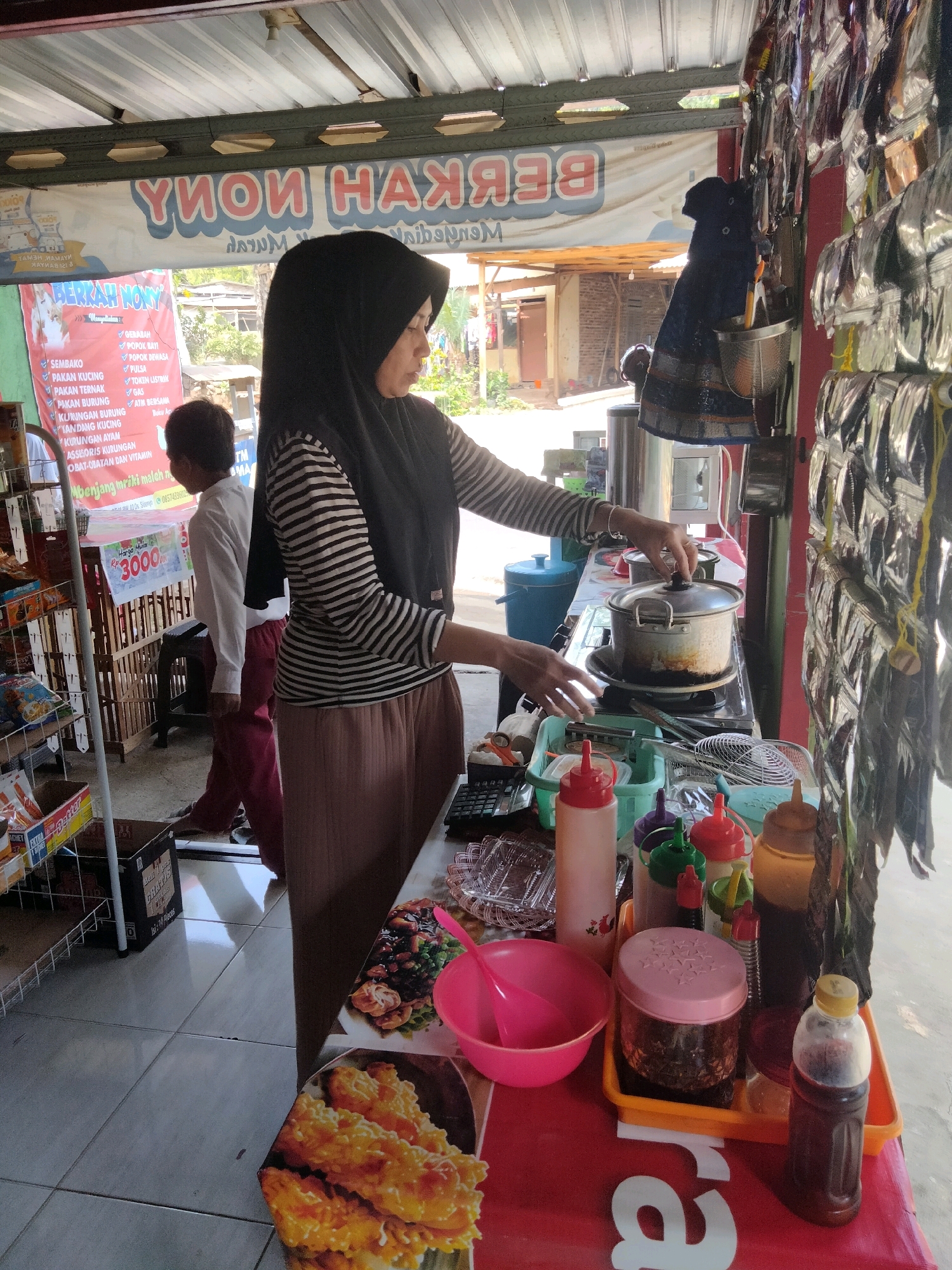The image shows an outdoor food stall in Sidorejo, Indonesia. A woman wearing a black hijab and a striped shirt is cooking on a portable stove. She is stirring a pot on the stove. In front of her is a table with various food items and condiments, including bottles of sauce, a pink bowl, and what appears to be fried food on a plate. To her left, a young boy in a white shirt and maroon pants is standing. Behind the woman, shelves are stocked with various goods, including what appear to be packaged snacks and drinks. A banner with Indonesian text is visible on the stall. The overall scene depicts a busy street food vendor preparing food.