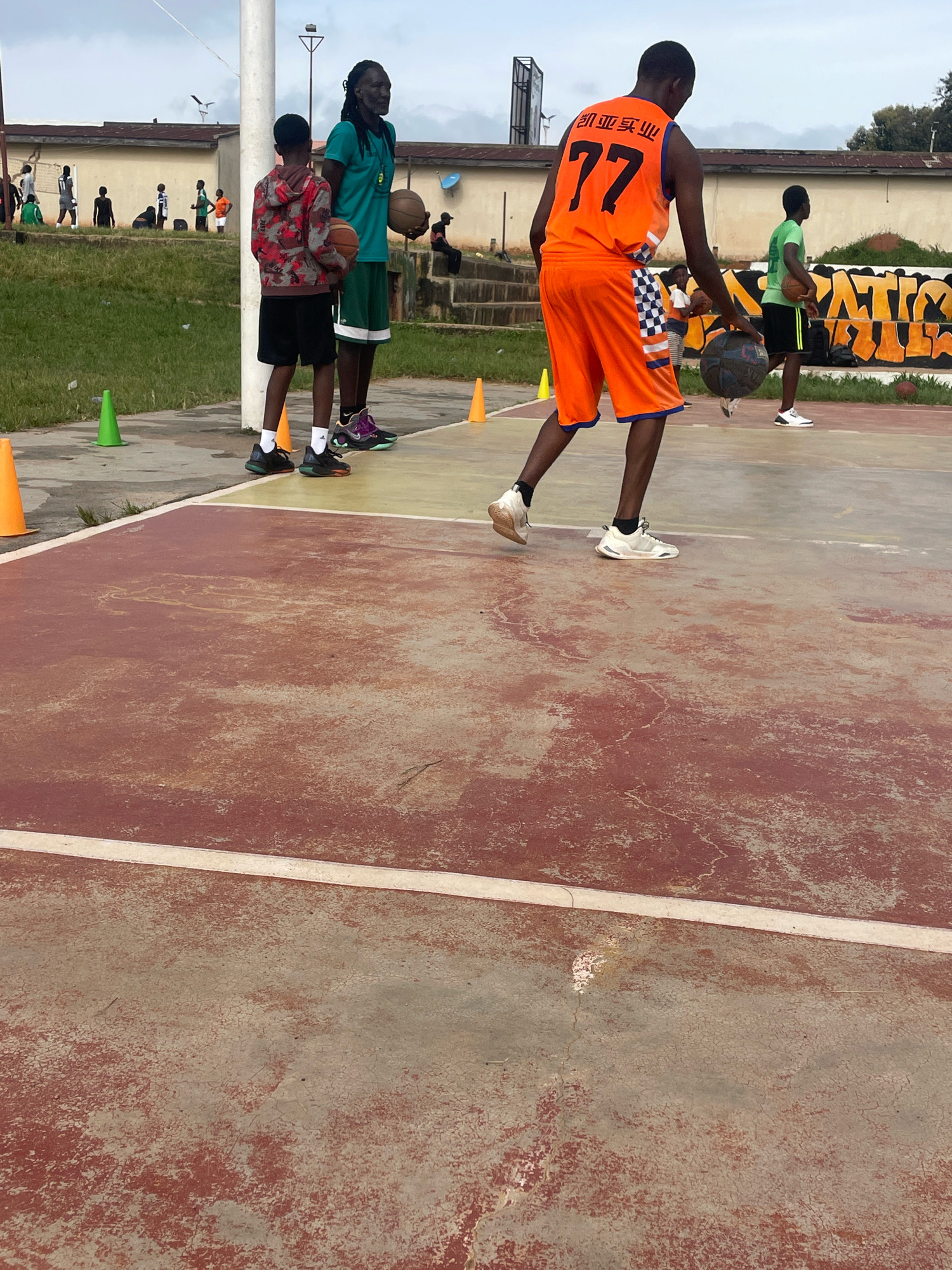In Gura Top, Nigeria, a group of individuals are engaged in a basketball game on an outdoor court. The court surface is weathered red concrete with yellow painted lines. The sky above is overcast. In the foreground, a young person in an orange basketball jersey with the number 77 prominently displayed and orange shorts is dribbling a dark basketball. To their left, another young person wearing a red and grey camouflage hoodie and black shorts stands holding a basketball. Further back, a tall individual with dark dreadlocks, wearing a teal t-shirt and green shorts, also holds a basketball. Another person in a green t-shirt and black shorts is visible in the mid-ground, holding a basketball. In the background, a low-rise building with a light-colored facade and a chain-link fence is present, along with some trees and a billboard. Several orange and green traffic cones are strategically placed on the court. The overall atmosphere suggests an informal basketball practice or game.