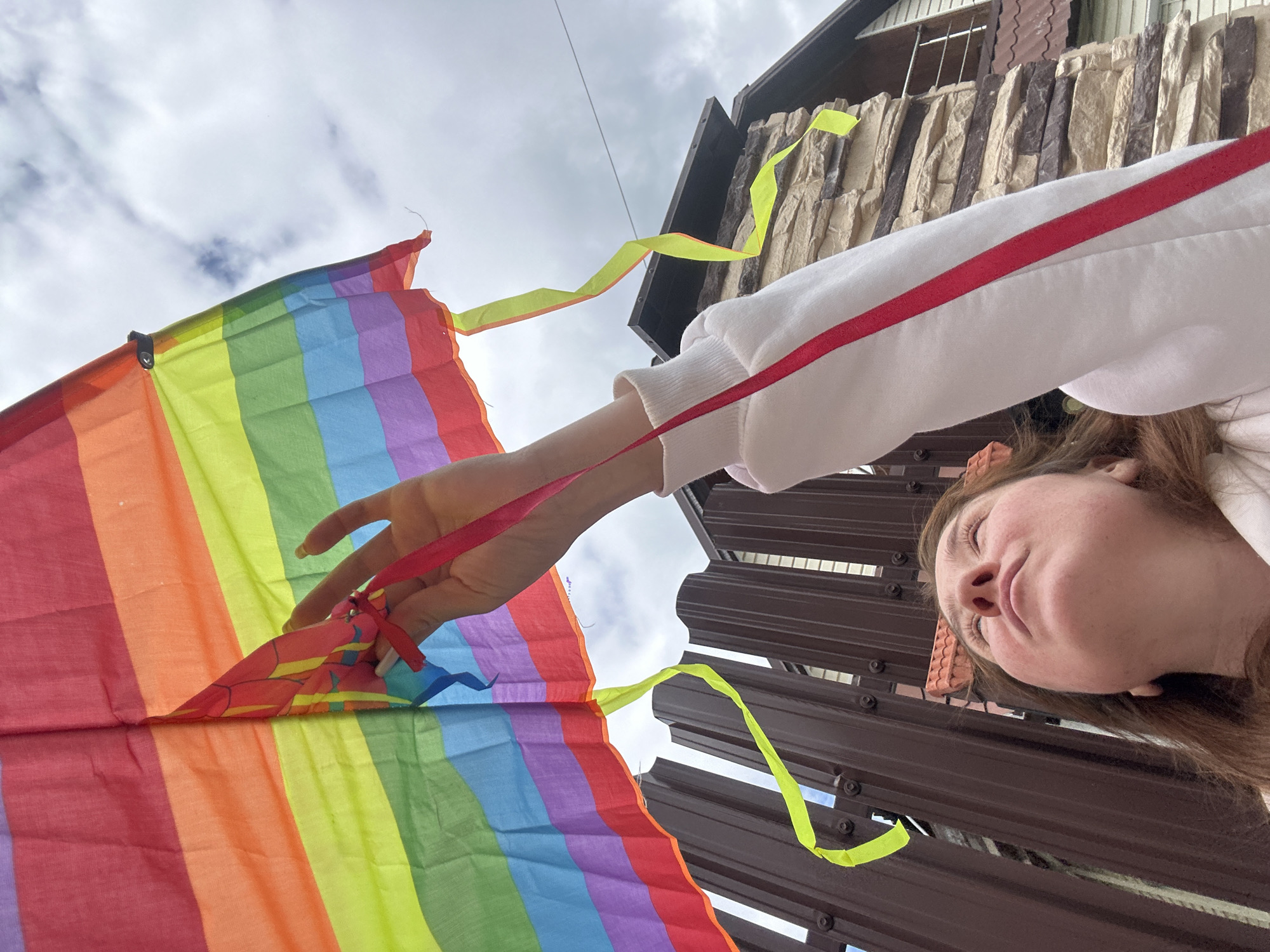 A young woman is shown from a low angle, looking up at a rainbow-colored kite. She is wearing a white sweatshirt with a red stripe down the sleeve. The kite is held by her outstretched arm, with strings and streamers visible. Behind her, a brown metal fence and a stone-clad house are partially visible. The sky is overcast with clouds. The scene is set outdoors, likely in a residential area.