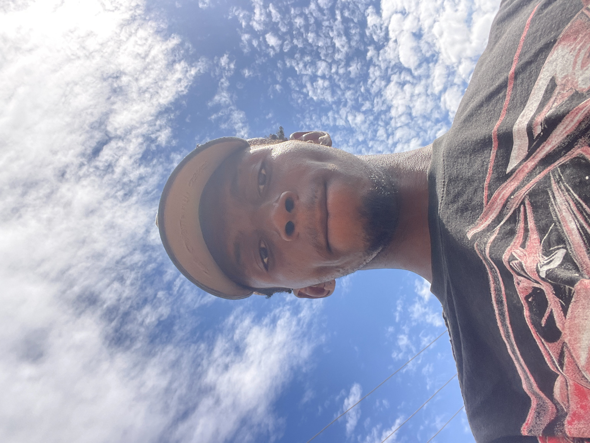 A young man takes a low-angle selfie, looking upwards into a bright blue sky filled with scattered white clouds. His face, visible from the nose down, occupies the center-left of the frame. He has a light beard or stubble and wears a light-colored baseball cap, the brim of which is prominent on the left. On the right, his dark t-shirt with red and white graphic designs is visible. The background is dominated by a clear, vibrant blue sky punctuated by numerous small, fluffy white clouds, suggesting a sunny daytime. Faint power lines are discernible against the sky in the lower right. The man's expression is neutral. While the context places the image in Lokoja, Nigeria, no specific geographical markers are present in the photo itself. No discernible text is clearly visible on his attire.