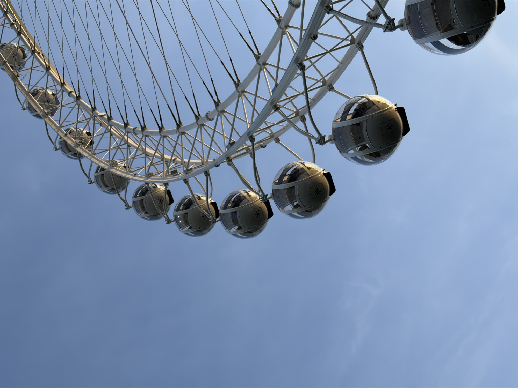 The image captures a close-up, low-angle view of a Ferris wheel against a clear blue sky. The Ferris wheel's structure is a complex network of white beams and thin cables, supporting multiple enclosed pods that hang from the outer rim. Several pods are visible, appearing as rounded, glass-enclosed cabins with some internal lighting. One pod in the foreground has a prominent logo or symbol on its exterior, resembling a stylized star or flower. The scene is dominated by the mechanical structure of the Ferris wheel and the vast expanse of the sky, with no people or specific city landmarks directly identifiable within the frame. Based on the typical appearance of this Ferris wheel design, it is likely the "Eye of St. Petersburg" or a similar amusement ride located in the city.
