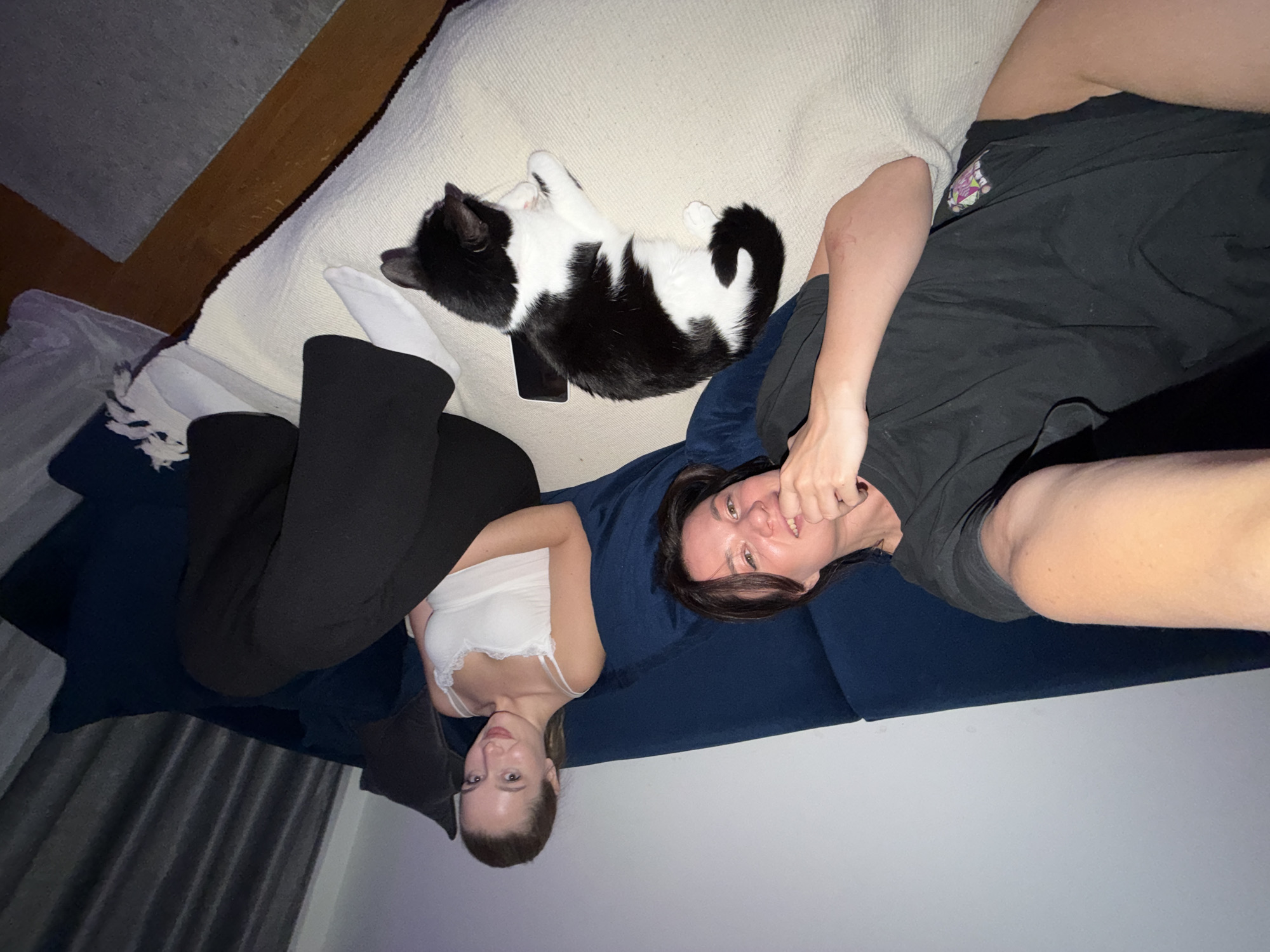 Two people and a cat are lying on a sofa. The first person is on the left, wearing a white top and black pants. The second person is on the right, wearing a black t-shirt and black shorts. The cat is black and white and lying on a white cushion next to the first person. A phone is near the cat. The sofa is blue. The room appears to be indoors. The location is not identifiable as being in Belgrade, Serbia.