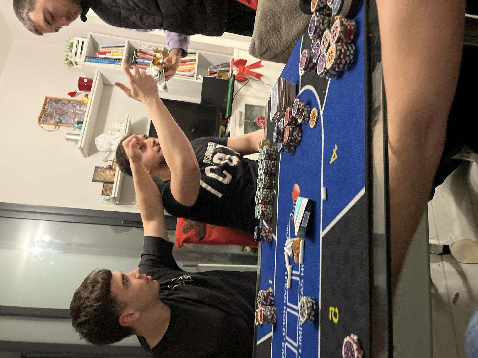 The image shows a group of people playing poker around a poker table. The table is covered in a blue felt with poker chips, cards, and other poker-related items on it. Two men are standing by the poker table. One man has his arms raised in the air. The image appears to be taken indoors in someone's home. The exact location in Burgas, Bulgaria, cannot be determined from the image.