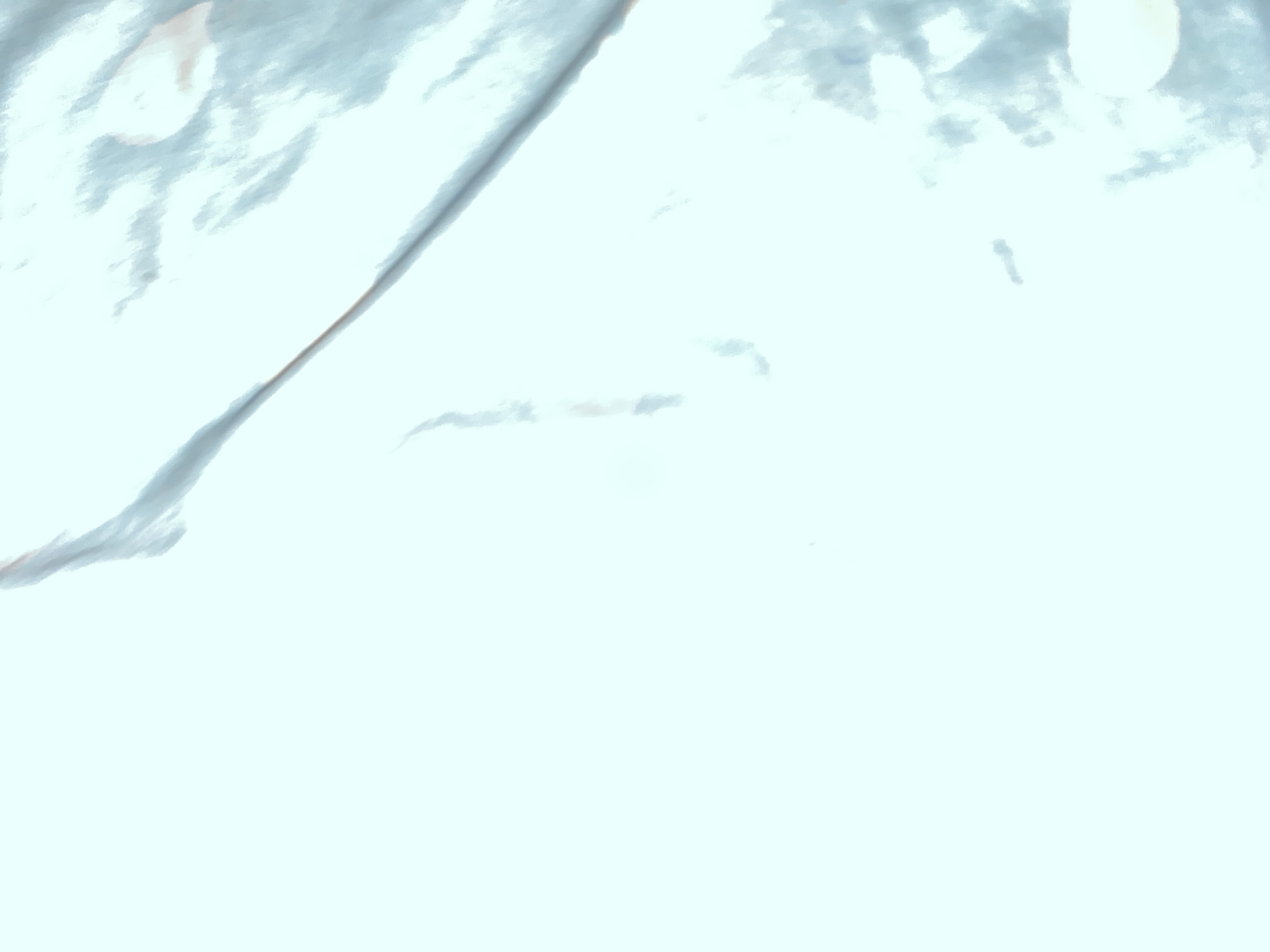 This is an abstract, digitally rendered image with a pale blue and white color palette. The composition features a large, amorphous shape with a textured, somewhat metallic or silky appearance dominating the upper left portion of the frame. This shape is characterized by soft, blended lines and subtle variations in light and shadow, suggesting a creased or folded material. A thin, darker line runs diagonally across this area, adding a subtle linear element. The rest of the image is a smooth, gradient of white and very pale blue, with a few indistinct, blurry shapes and specks scattered across the lower and right portions. These smaller elements appear as smudges or faint suggestions of form, adding a sense of depth or texture to the otherwise blank background. There is no discernible narrative, recognizable objects, or human figures. The overall impression is one of soft light and abstract texture, evoking a sense of calmness or perhaps digital noise.
