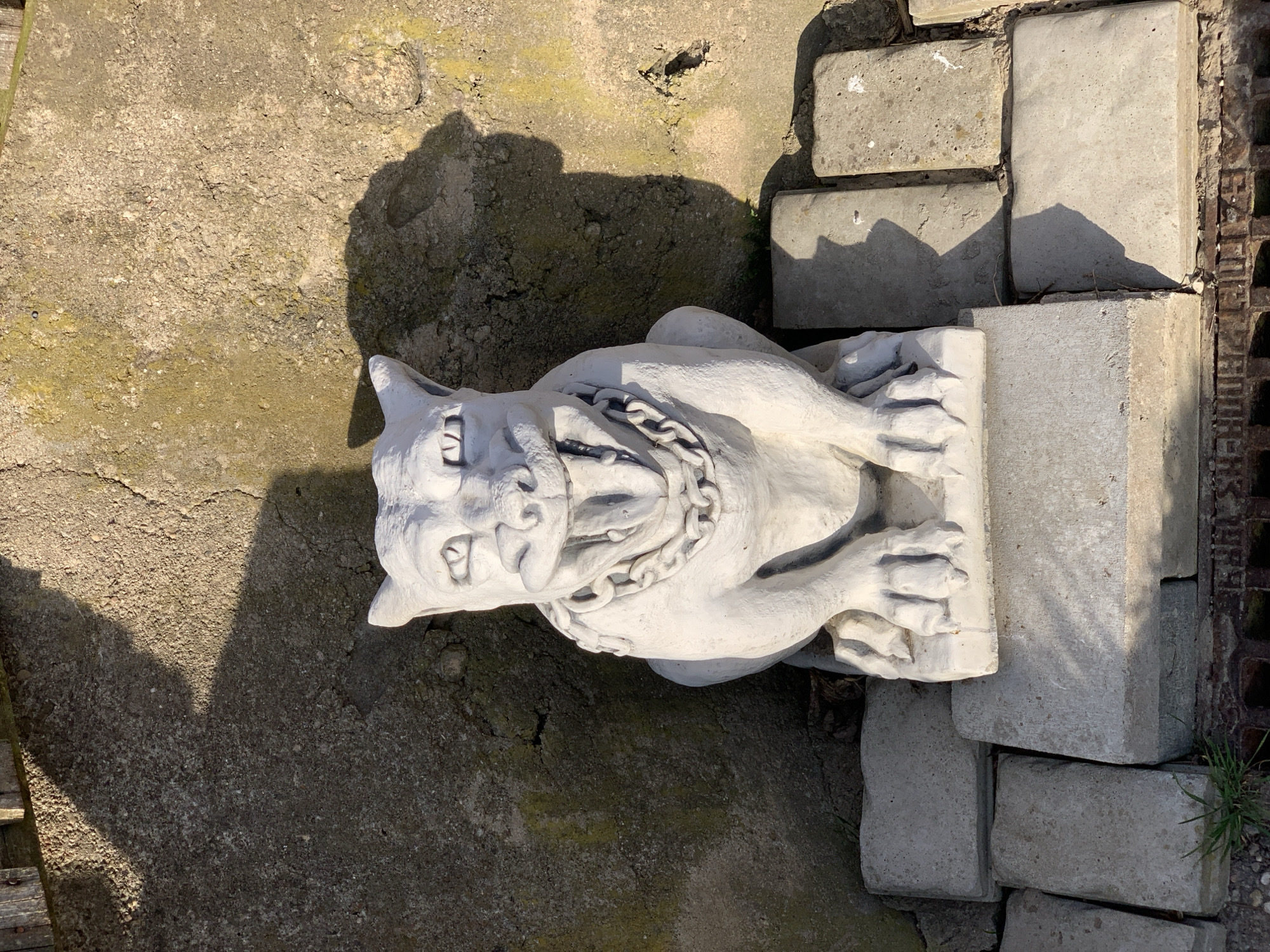 The image depicts a white, concrete statue of a snarling dog with a chain around its neck, lying on its side. The statue is positioned on a surface of cracked, gray concrete, with a pile of rectangular, concrete blocks stacked to the right of the statue. A section of rusty metal grating is visible along the far right edge. The location appears to be an outdoor, possibly unkempt, area. The geographical location in Hrodna, Belarus cannot be inferred solely from the image.
