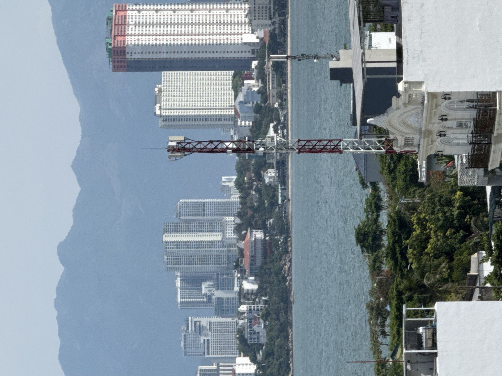 This media file presents a panoramic view of a coastal city, likely located in Bac Nha Trang Ward, Vietnam. The image is captured from an elevated perspective, possibly from a building or a drone, looking out over a large body of water.
The scene is dominated by a dense collection of high-rise buildings, characteristic of an urban coastal environment. These structures vary in architectural style and color, with many featuring predominantly white facades and numerous windows, suggesting residential or hotel complexes. Some buildings are taller and more modern, while others appear to be older or of a different design. Among the buildings, a prominent red and white construction crane is visible, hinting at ongoing development or infrastructure projects within the city.
The foreground includes parts of a modern white building with balconies and ornate architectural details, possibly a hotel or resort. Lush green trees and foliage are interspersed between the buildings and along the coastline, adding a touch of nature to the urban landscape. The body of water, likely the sea, stretches across the frame, appearing calm with gentle ripples. In the background, a hazy mountain range can be observed, providing a natural backdrop to the city.
The lighting suggests it is daytime, with a bright, diffused light indicating possibly overcast or slightly hazy weather, common in coastal regions. There are no visible people or specific activities taking place that can be discerned from this distant and somewhat abstract perspective. The overall impression is one of a bustling, developing city situated by the sea, with a blend of modern infrastructure and natural scenery. No visible text is present in the image.