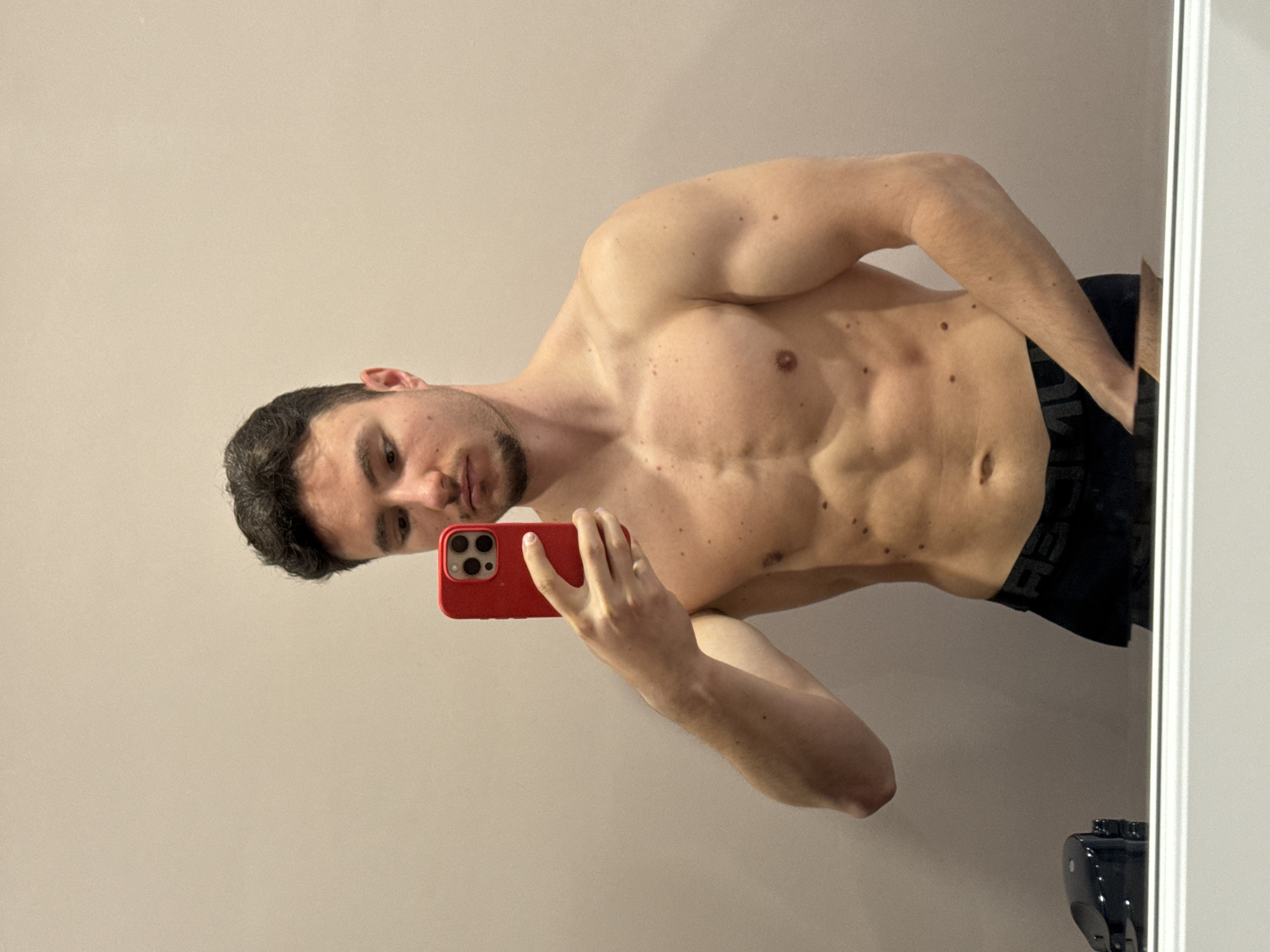 The media file presents a mirror selfie of a shirtless young man, captured indoors. He exhibits a muscular physique, with visible abdominal muscles, defined chest, and arms. His short, dark hair and light facial hair (stubble/short beard) are visible. He holds a red smartphone with multiple camera lenses, using it to take the picture, and his eyes are directed towards the camera.
Numerous moles are present on his torso and arms. He is wearing black underwear or shorts, with the partial text "UNDER" discernible on the waistband. The background is a plain, light-colored wall, reflecting the indoor environment. The image itself is rotated, suggesting the photo was taken in a landscape orientation. In the bottom right corner, a small, dark object is partially visible behind the white frame of the mirror. The lighting is even and soft, indicative of indoor conditions. The scene suggests a casual self-portrait.