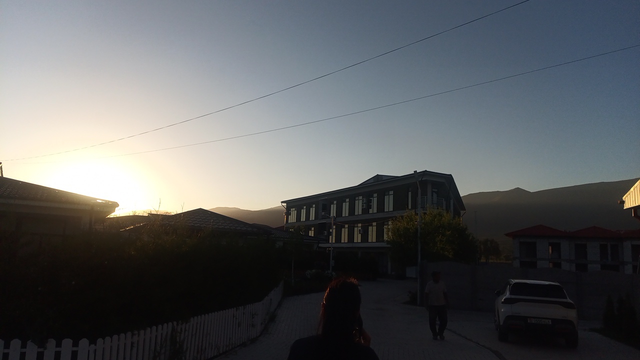 The image shows a street scene in Choktal, Kyrgyzstan, at dusk. A person's back is to the camera, with their head turned to their right, appearing to be on their phone. They are walking on a paved road. To the left, a white picket fence runs along the side of the road, with dark foliage behind it. A house is visible beyond the fence. In the center of the image, a three-story building with numerous windows is situated. To the right, a white SUV is parked, facing away from the camera. Further in the background, mountains are silhouetted against the sky. The sky is clear and transitioning from the bright yellow of sunset to a pale blue. Two power lines are visible stretching across the sky.