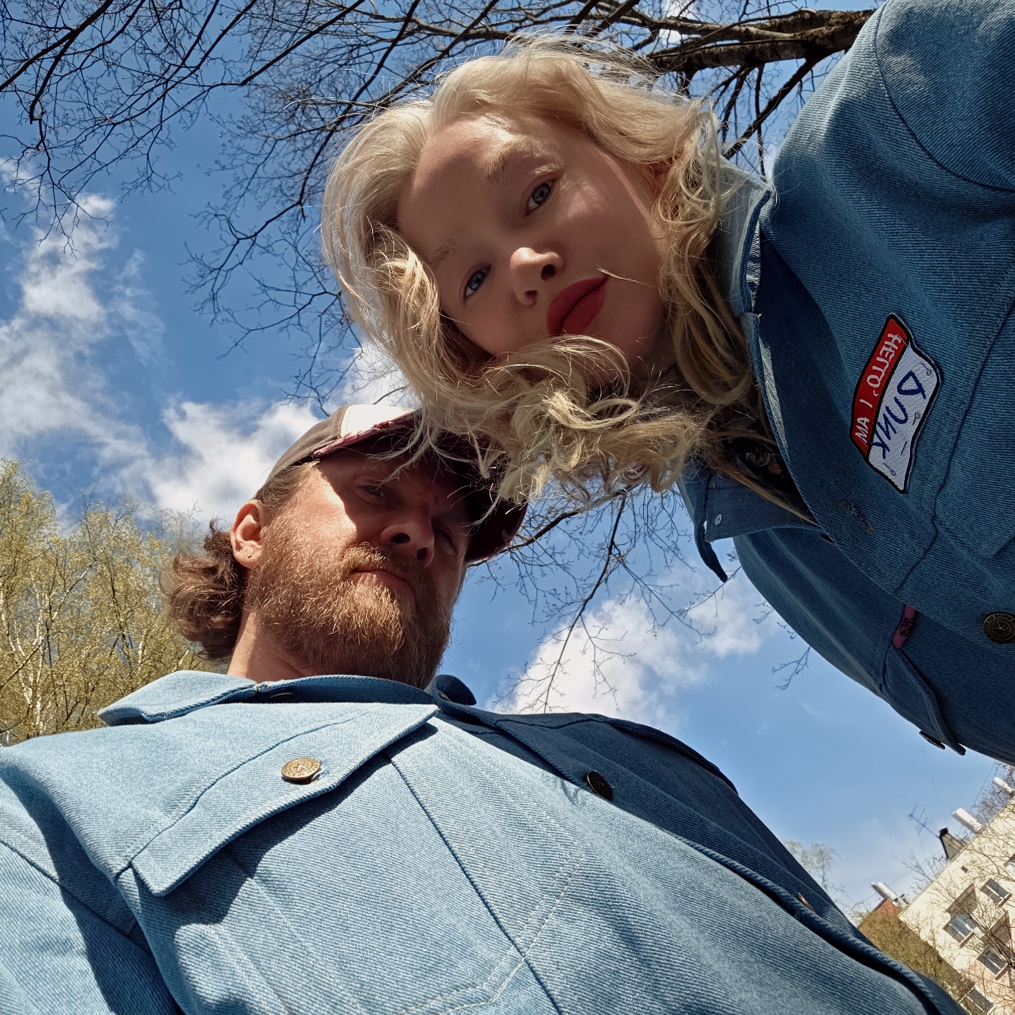 A low-angle outdoor portrait features two individuals, a woman and a man, looking directly down at the camera from above. The woman, positioned in the upper right, has long, wavy blonde hair, blue eyes, and red lipstick, wearing a light blue denim jacket. On her right shoulder is a red and white patch with mirrored text, partially visible as "6ЛИК" (BLIK) and "HETTO" (NETTO). The man, to her left and below, has a reddish-brown beard, a brown and white baseball cap, and is also wearing a light blue denim jacket or shirt. His expression is stoic.
The background consists of a bright, clear blue sky with scattered white clouds, suggesting a sunny daytime. Bare tree branches are silhouetted against the sky in the upper left, while some trees with light green, budding leaves are visible in the lower left. In the far distance on the lower right, glimpses of light-colored urban buildings with dark roofs can be seen, indicating an urban or suburban setting, likely in Saint Petersburg, Russia. The strong natural light illuminates their faces and the textures of their denim attire. The overall impression is a casual, direct gaze captured from a unique perspective.