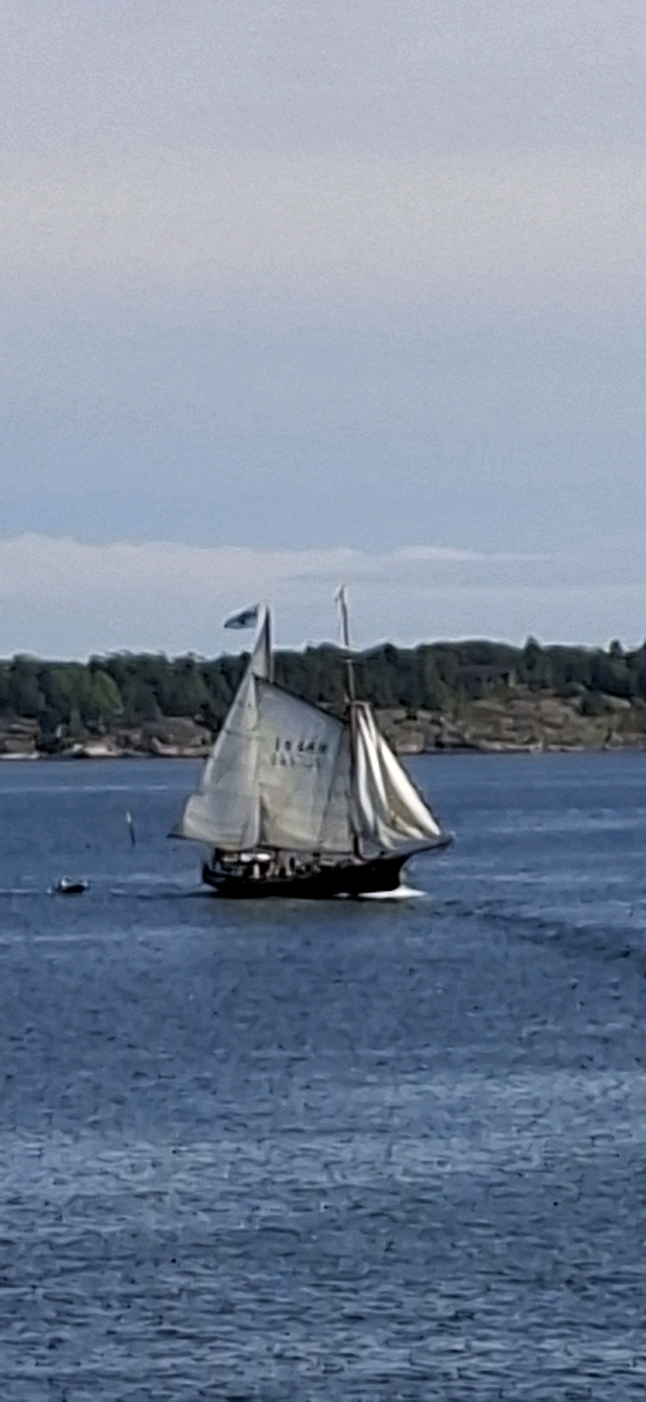 A large, two-masted sailboat with white sails is sailing on a body of water, likely the Baltic Sea. The boat has a dark hull and appears to have people on deck. The sails are filled with wind, indicating the boat is moving. In the background, a tree-covered island or coastline is visible under a cloudy sky. The water is a deep blue with some chop. A smaller boat is visible to the left of the sailboat. The image appears to have been taken from a distance. The sailboat is identified with the text "IN LAN" written on its sails, possibly indicating its name or origin. This scene is consistent with the archipelago surrounding Helsinki, Finland, where numerous islands and a bustling maritime environment are characteristic features.