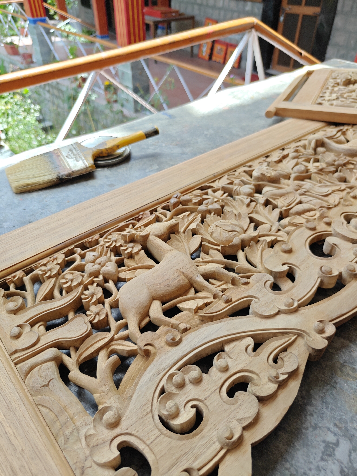 The image shows a close-up of an intricately carved wooden panel, likely a decorative element for a building or furniture. The carving depicts floral motifs and an animal, possibly a deer, amidst foliage. A wide paintbrush rests on a small metal container, suggesting the carving is being prepared for finishing or restoration. The panel sits on a grey surface, possibly stone or concrete, adjacent to a balustrade with a blurred background showing architectural elements like columns and a garden. The scene suggests a workshop or outdoor workspace in Fatehpur, India, where woodworking and carving are traditional crafts.